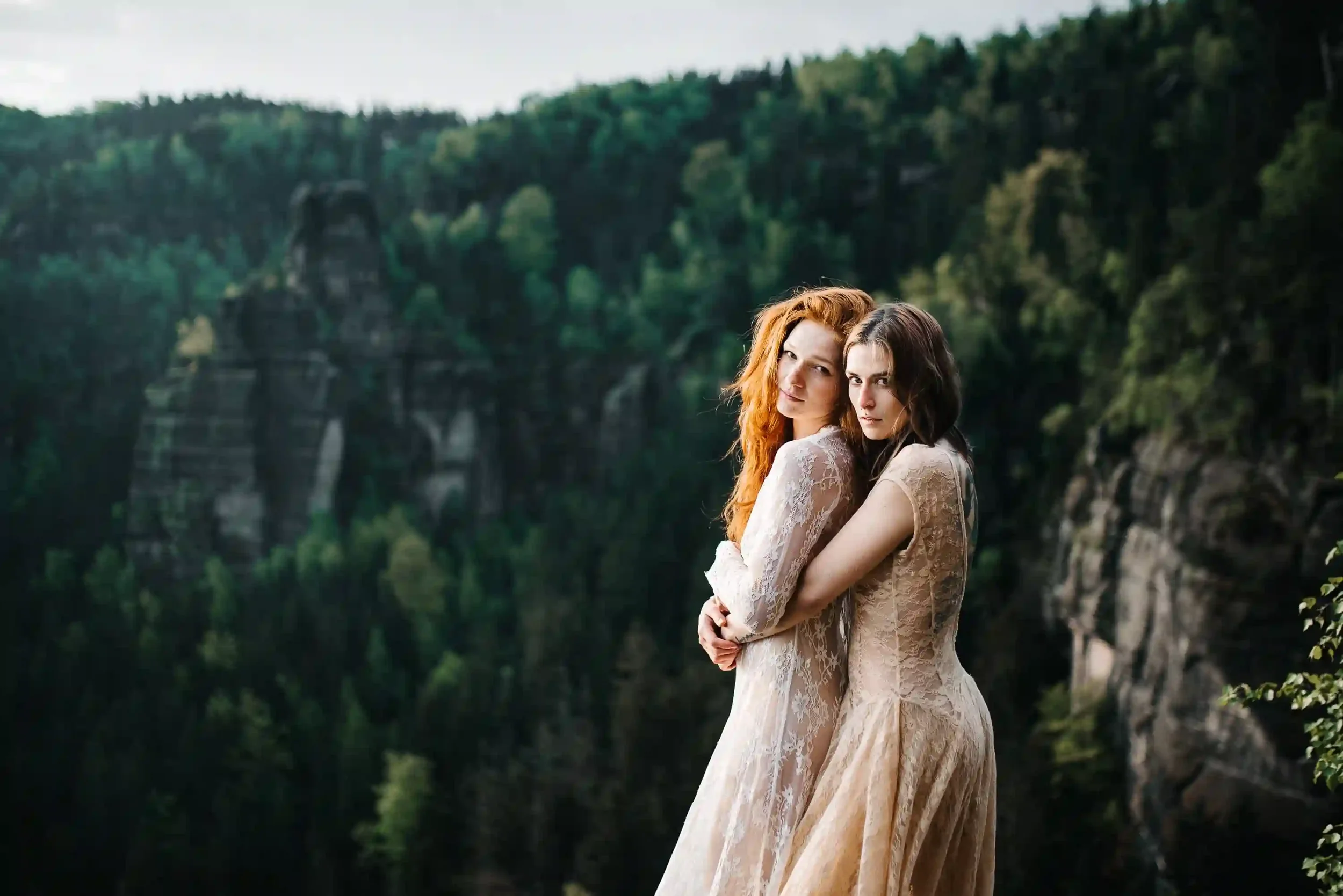 Two women in lace dresses stand close together, embracing and looking at the camera, with a forested canyon and rocky cliffs in the background.
