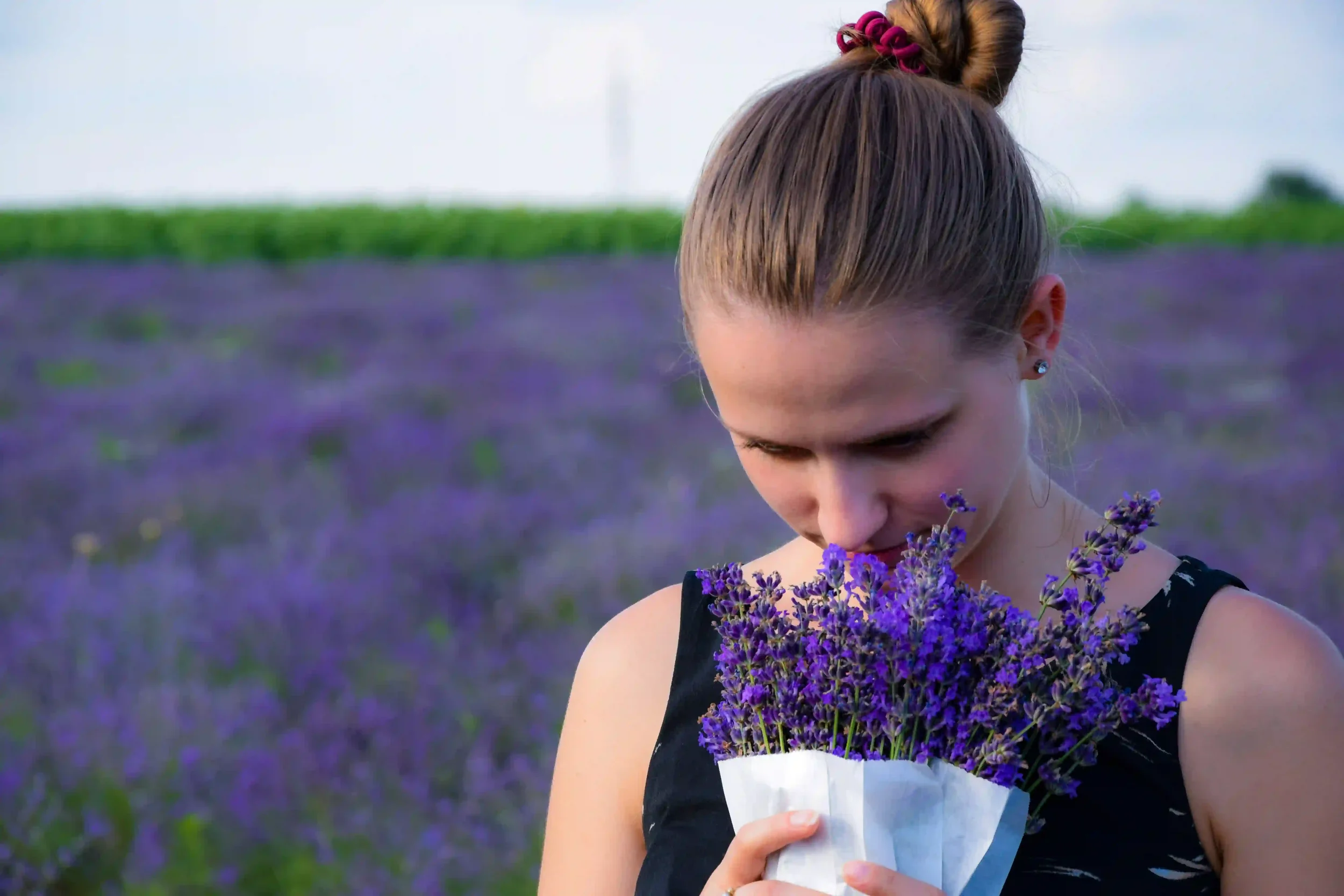 A young woman with blonde hair in a bun is standing in a lavender field, wearing a black sleeveless top, and smelling a bouquet of fresh lavender flowers wrapped in white paper.