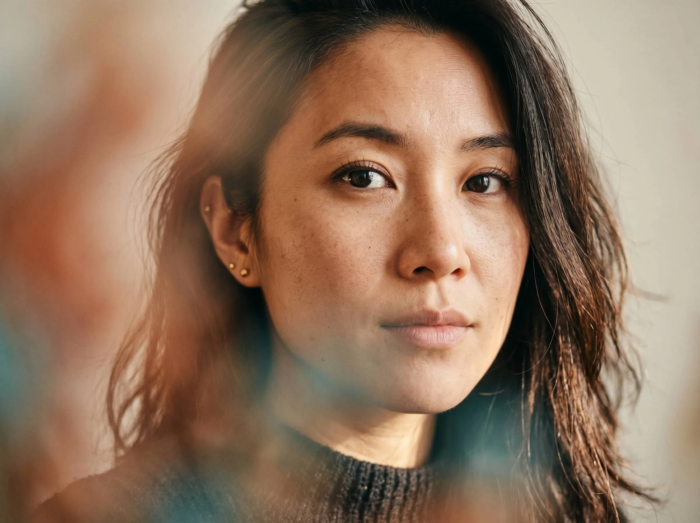 Close-up portrait of a woman with natural makeup, dark hair, and a neutral expression, set against a softly blurred background.
