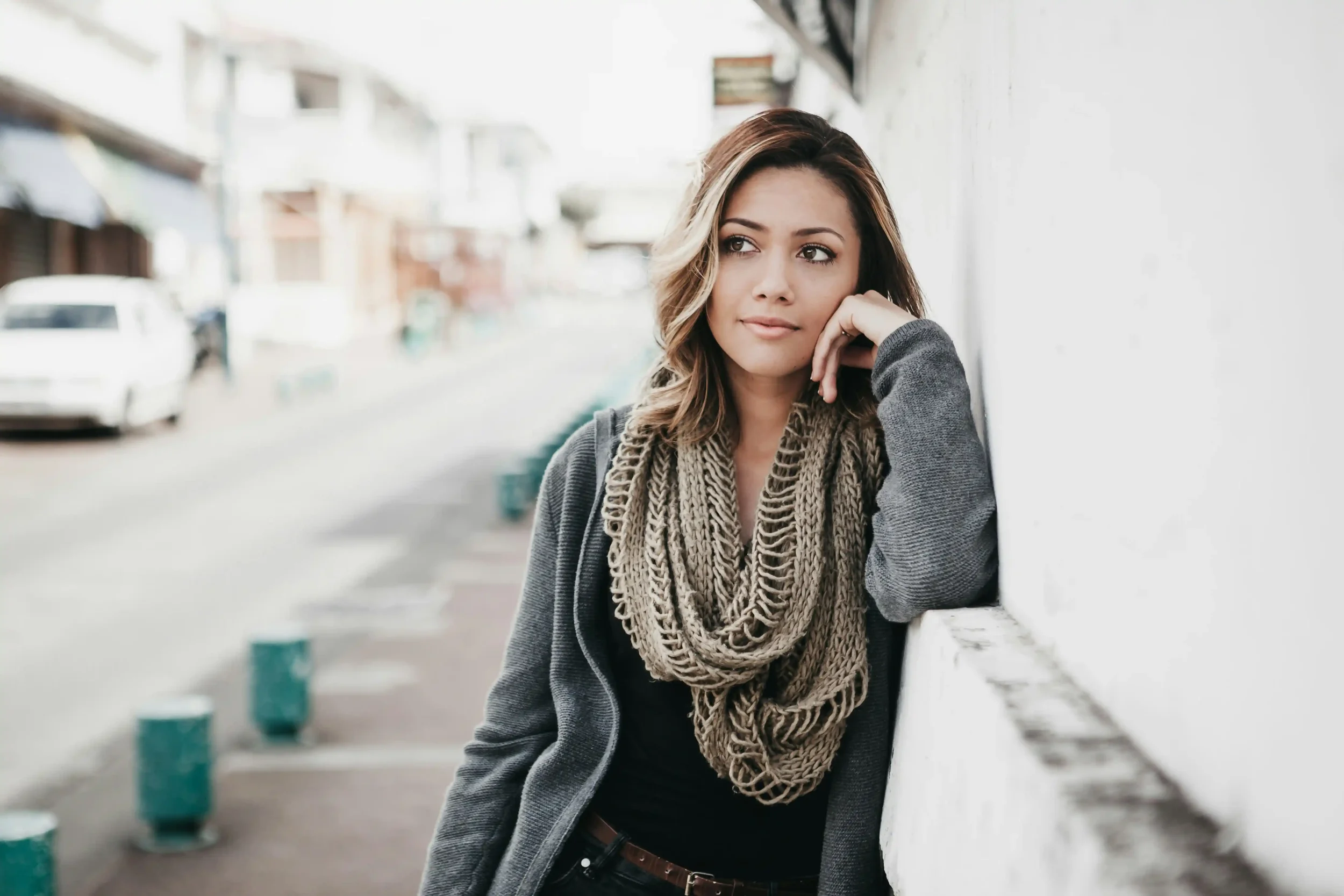 A young woman with wavy hair, wearing a chunky scarf and gray sweater, leans against a white wall on a quiet city street, gazing thoughtfully into the distance.