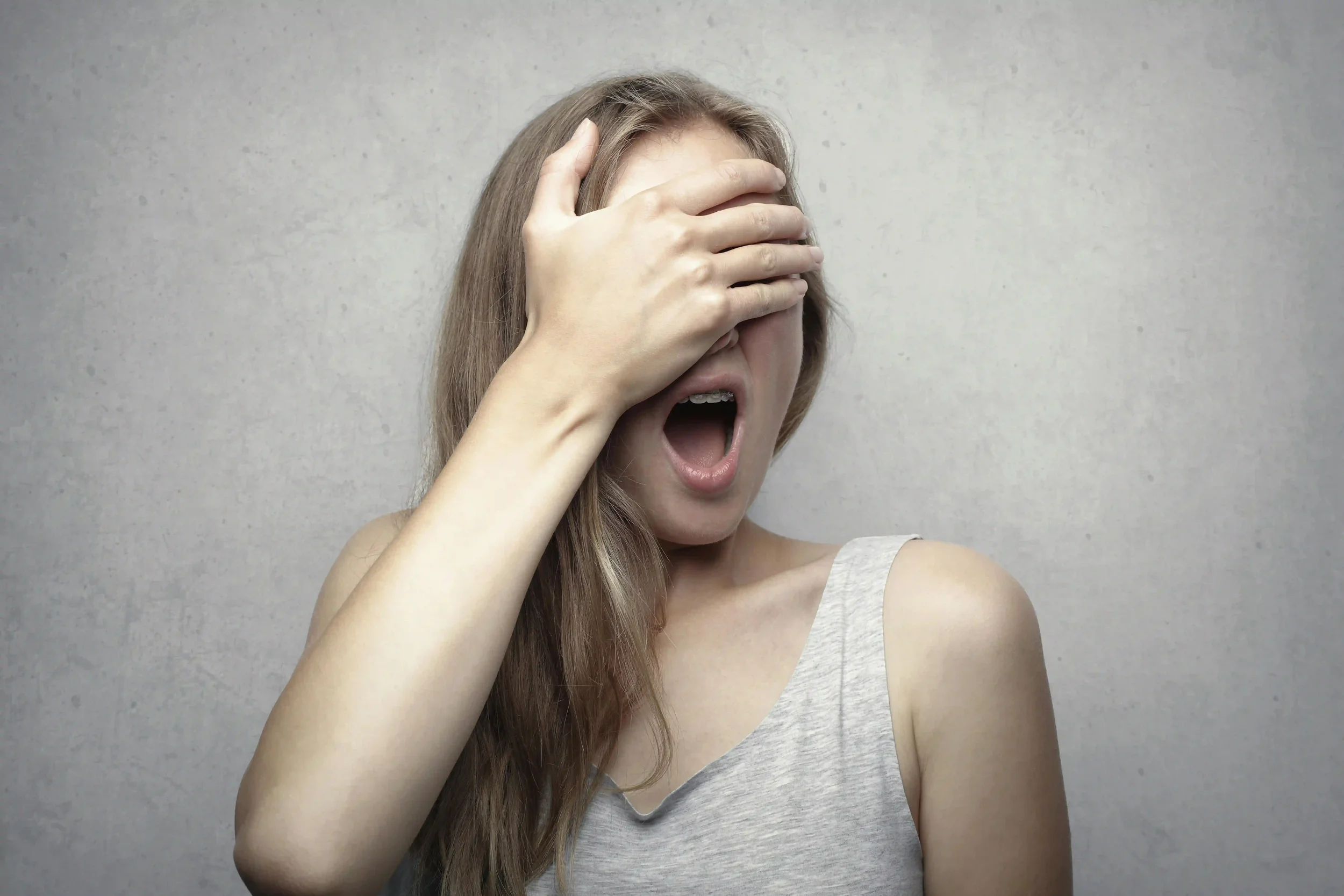 A woman with long hair covers her eyes with one hand while her mouth is open, as if surprised or shocked, against a plain gray background.