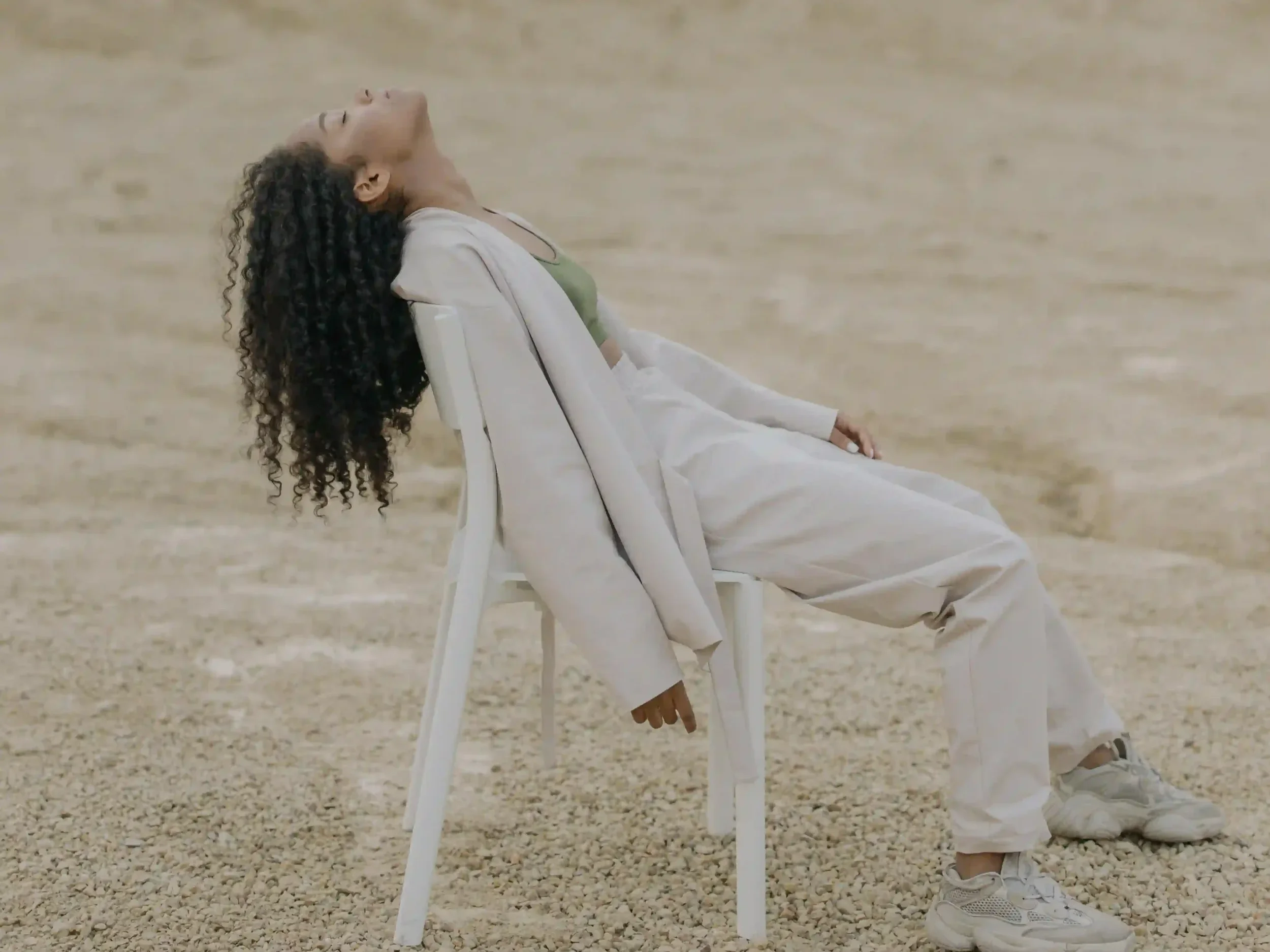 A woman with curly hair leaning their head and torso back while sitting on a white chair in a sandy landscape.