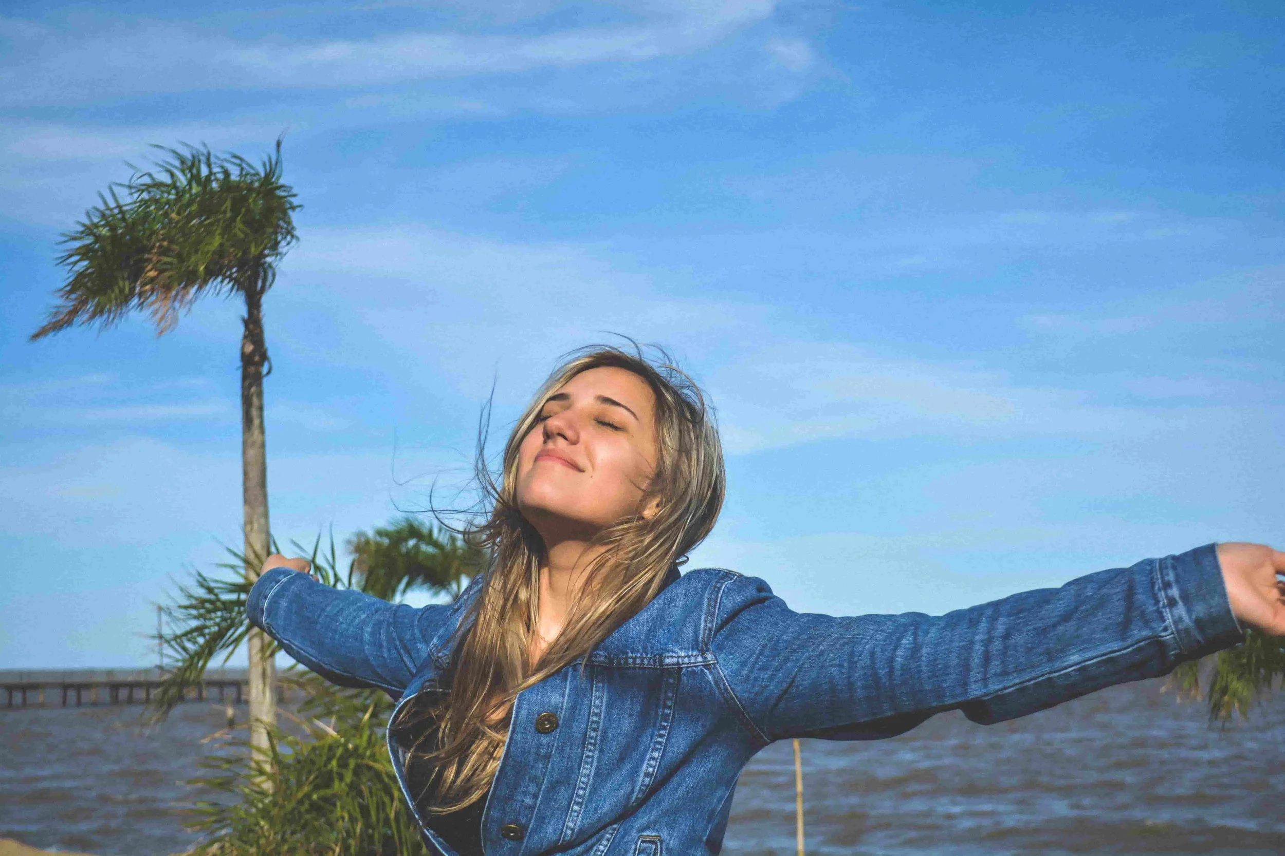 A young woman in a denim jacket stands outdoors with arms outstretched, eyes closed, and a peaceful smile. Palm trees and blue sky with wispy clouds are in the background, suggesting a breezy, sunny day by the water.