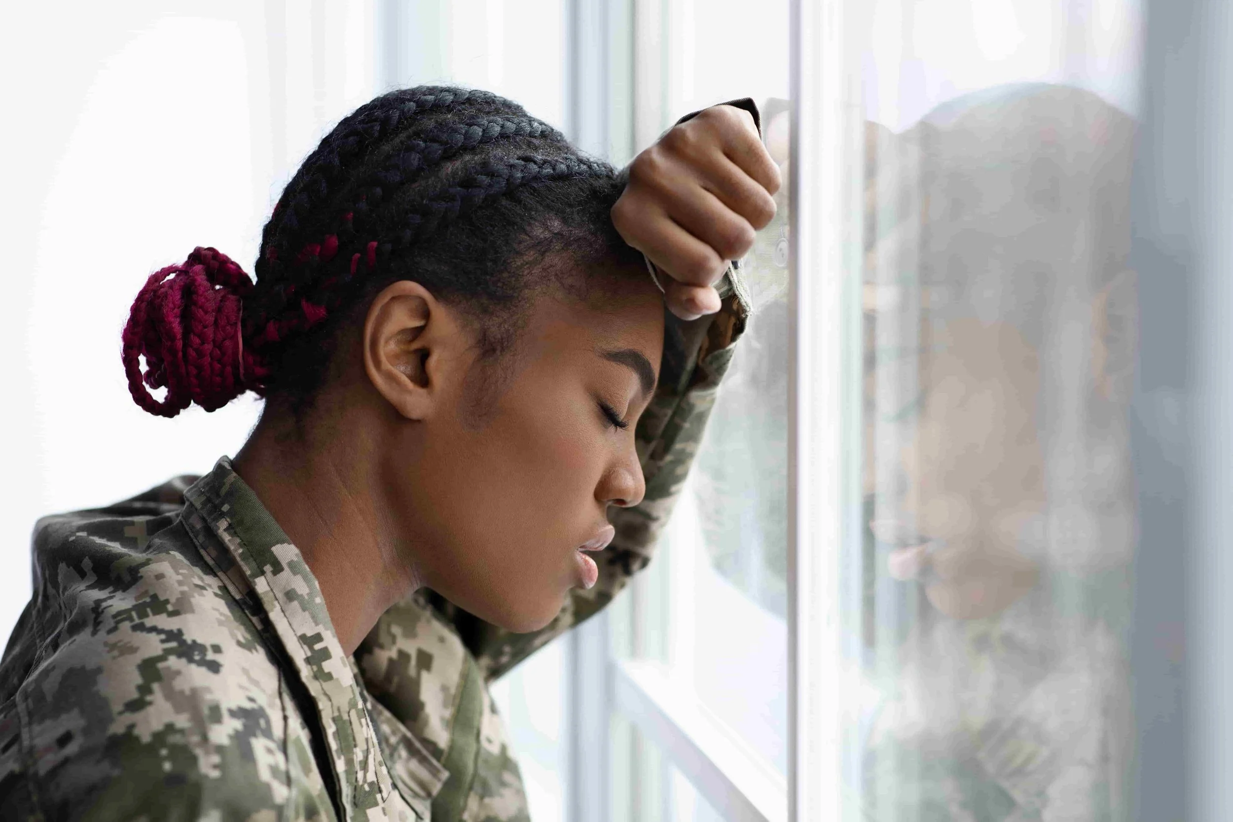 A woman in military uniform leans their forehead against a window, eyes closed and expression somber, suggesting feelings of stress or exhaustion.