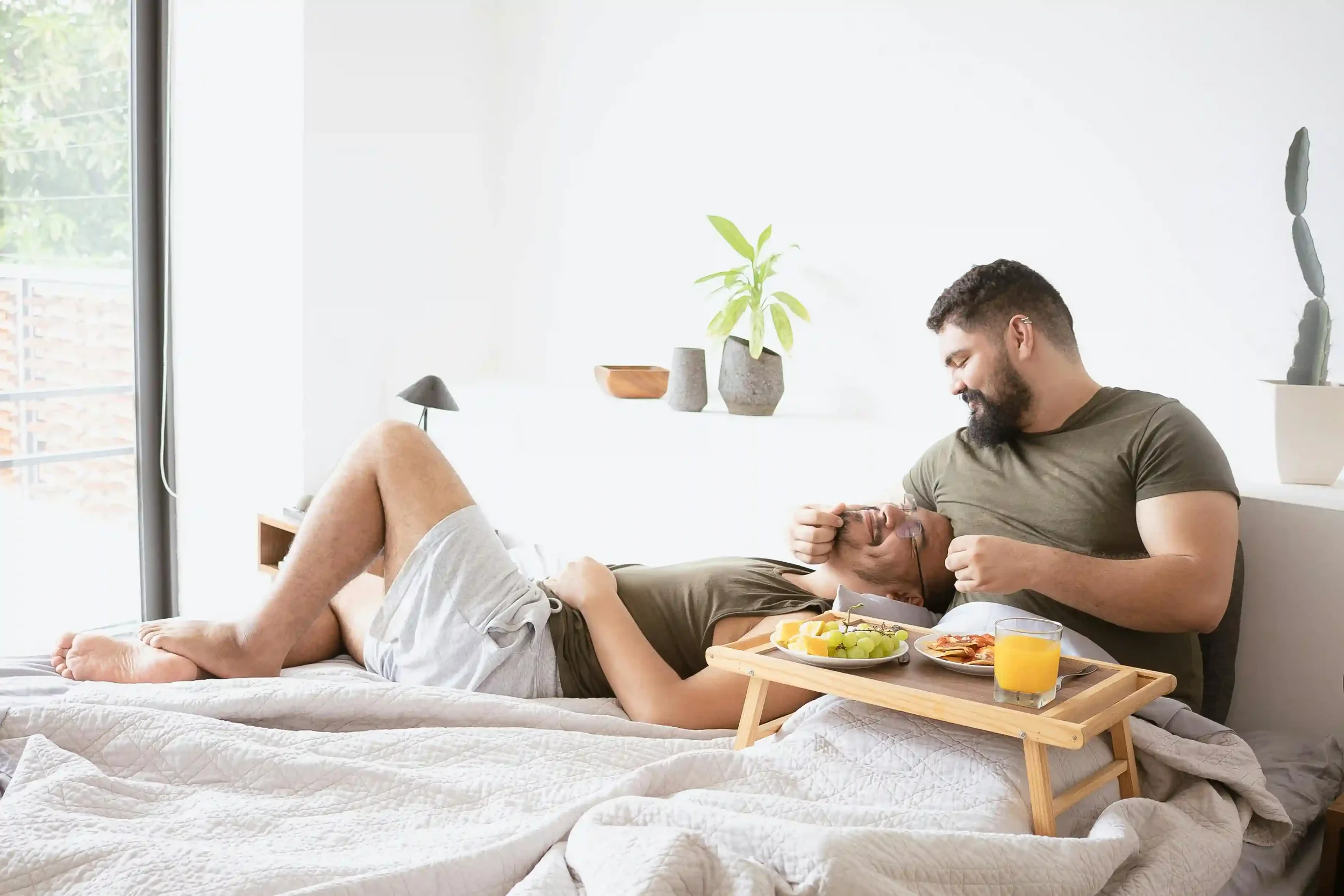 Two men relax on a bed together; one lies down with his head in the others lap. A breakfast tray with food and orange juice is in front of them, and they share a warm, affectionate moment in a bright bedroom.