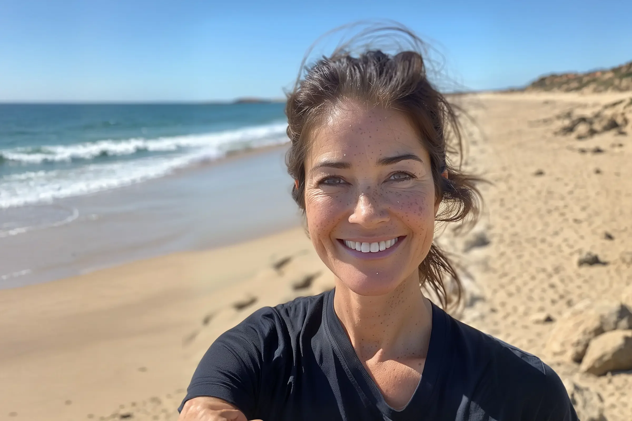A woman with brown hair and freckles smiles at the camera while taking a selfie on a sunny, sandy beach with gentle waves and clear blue sky in the background.