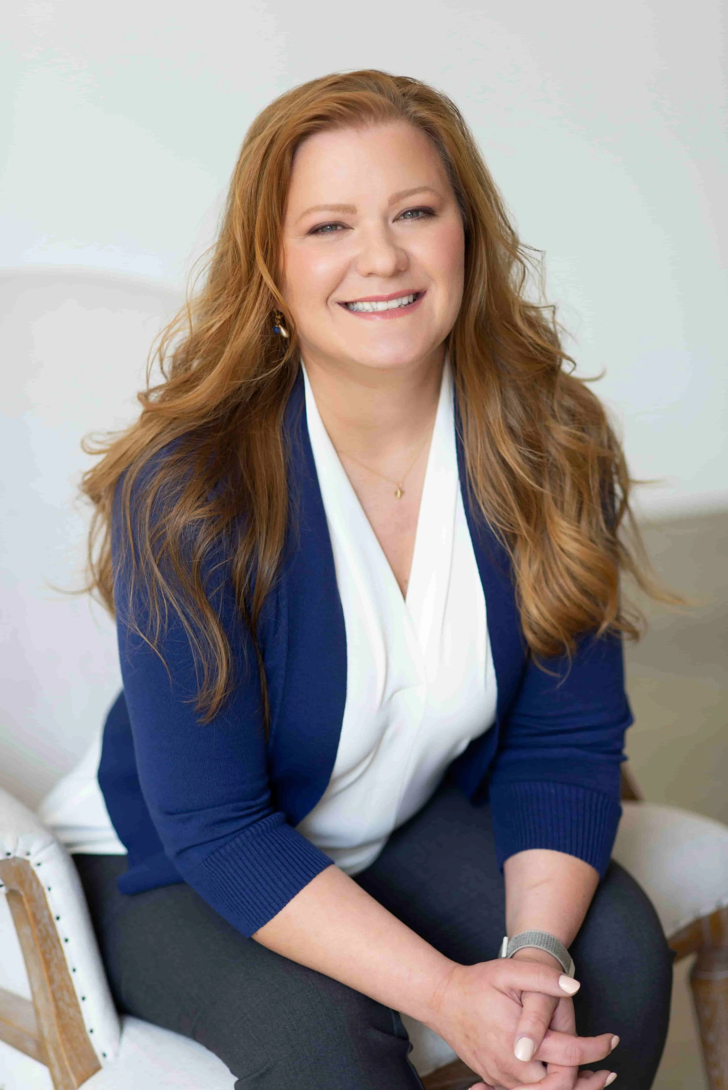 A picture of EMDR therapist Anna Khandrueva sitting on a light-colored chair, smiling warmly. Dressed in a white blouse and navy blue cardigan, she has her hands clasped in front of her as she provides therapy for women in Broomfield