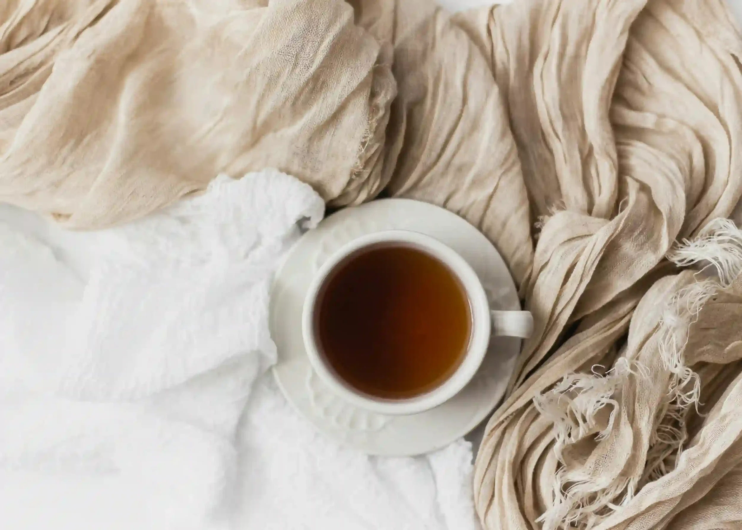 A white cup filled with tea sits on a matching saucer, surrounded by soft, beige and white textured fabrics, creating a cozy and inviting atmosphere.