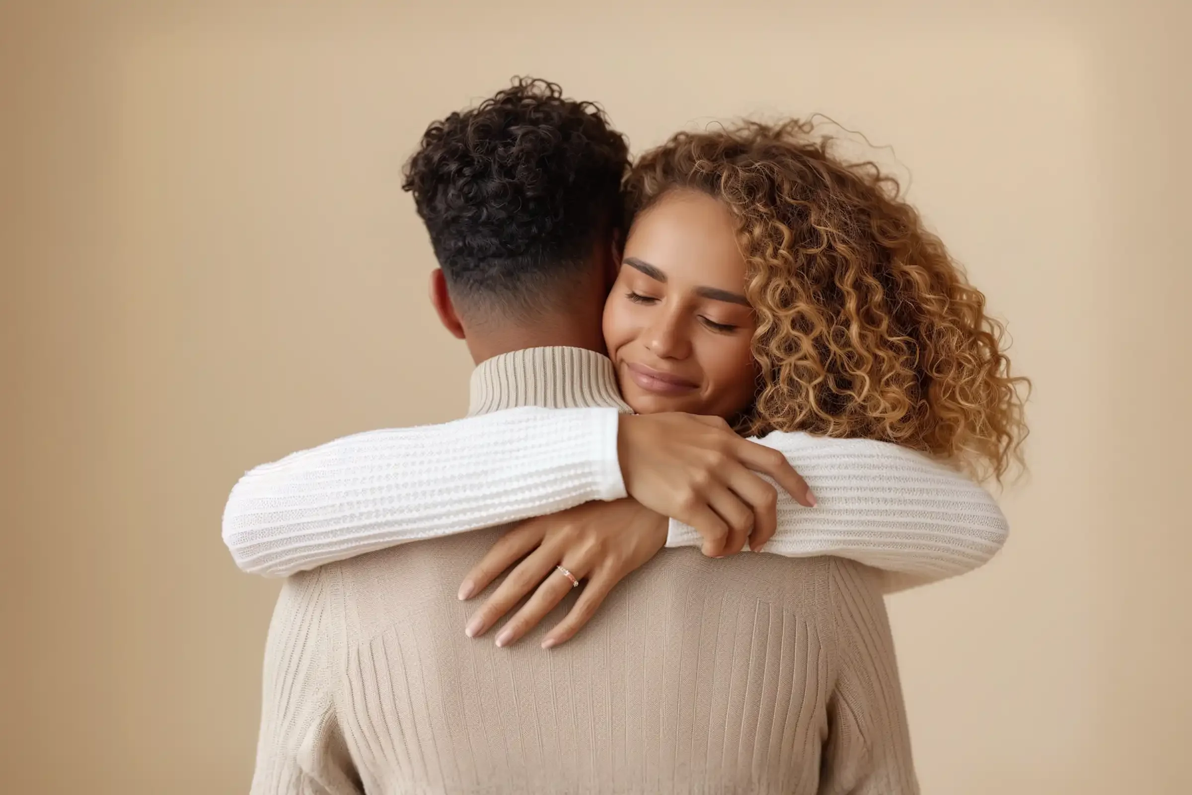 A woman with curly hair smiles with her eyes closed while hugging a man from behind. Both wear light-colored sweaters against a beige background, and her hand with a ring rests on his back.