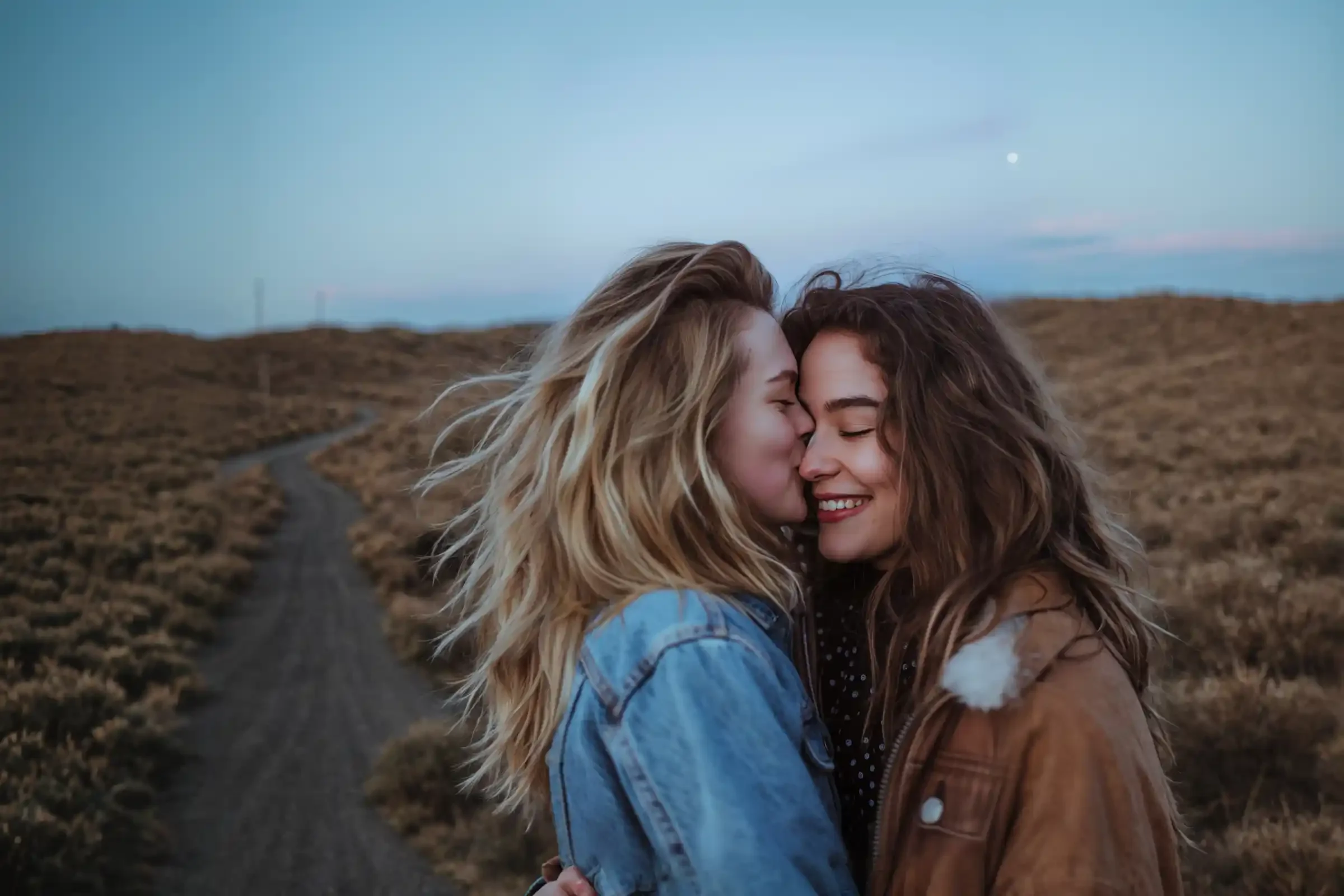 Two women embrace on a dirt path in a grassy field at dusk. One woman gently kisses the others forehead while both smile with closed eyes, creating an intimate and joyful moment.