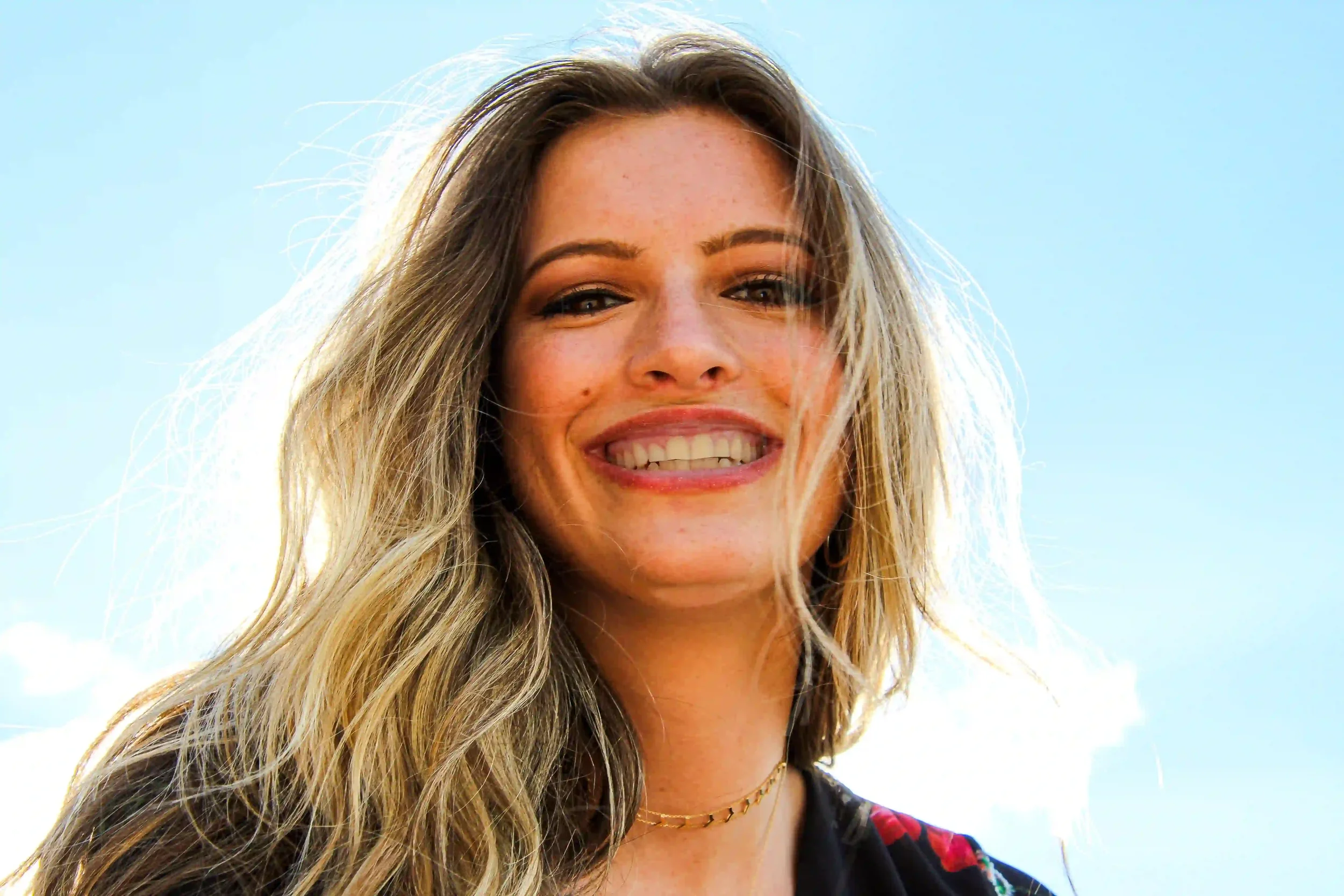 A young woman with long blonde hair smiles at the camera against a clear blue sky, wearing a black top and a necklace. Sunlight highlights her hair and face.