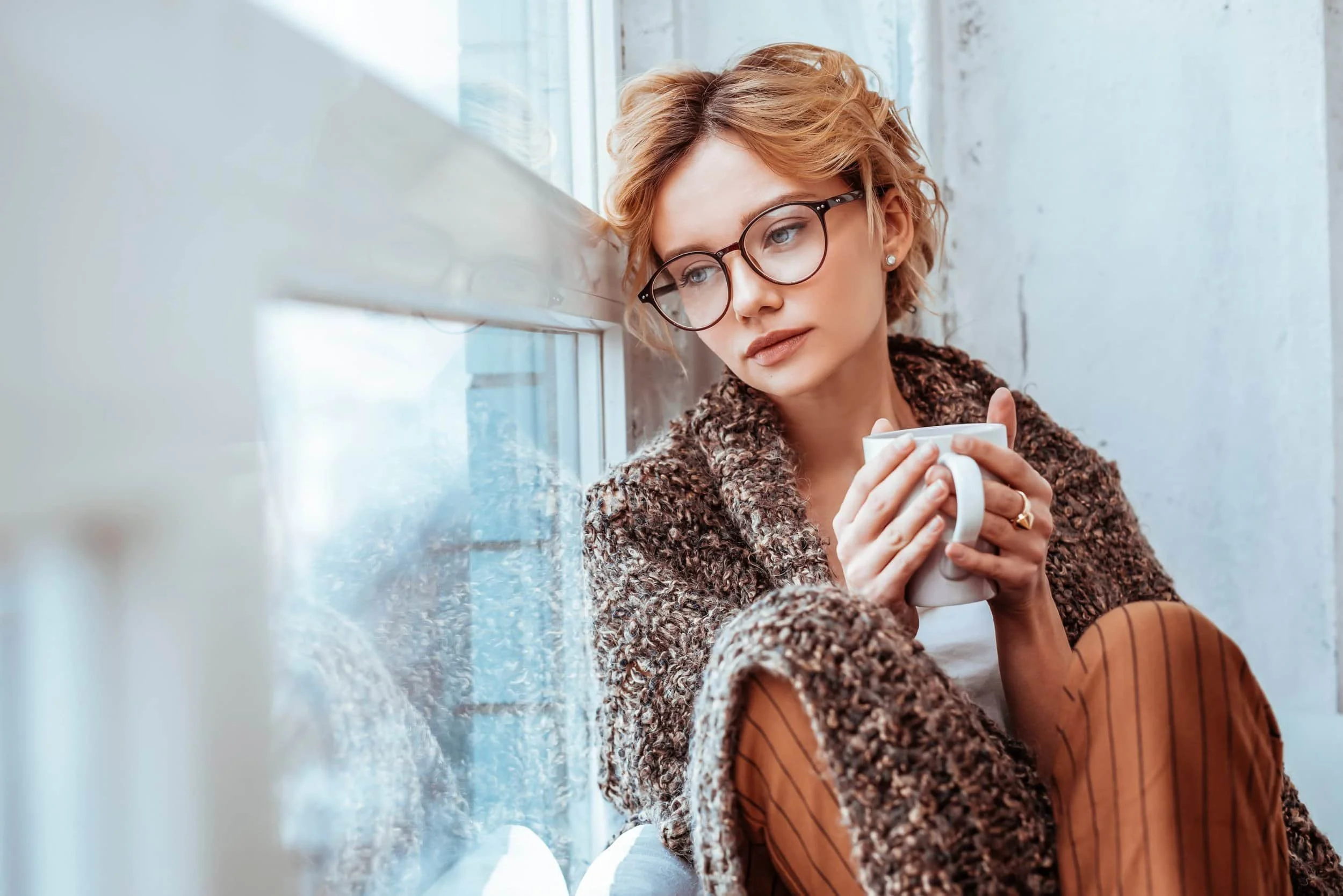 A woman with short blonde hair and glasses sits by a window, holding a white mug while wrapped in a knit shawl.