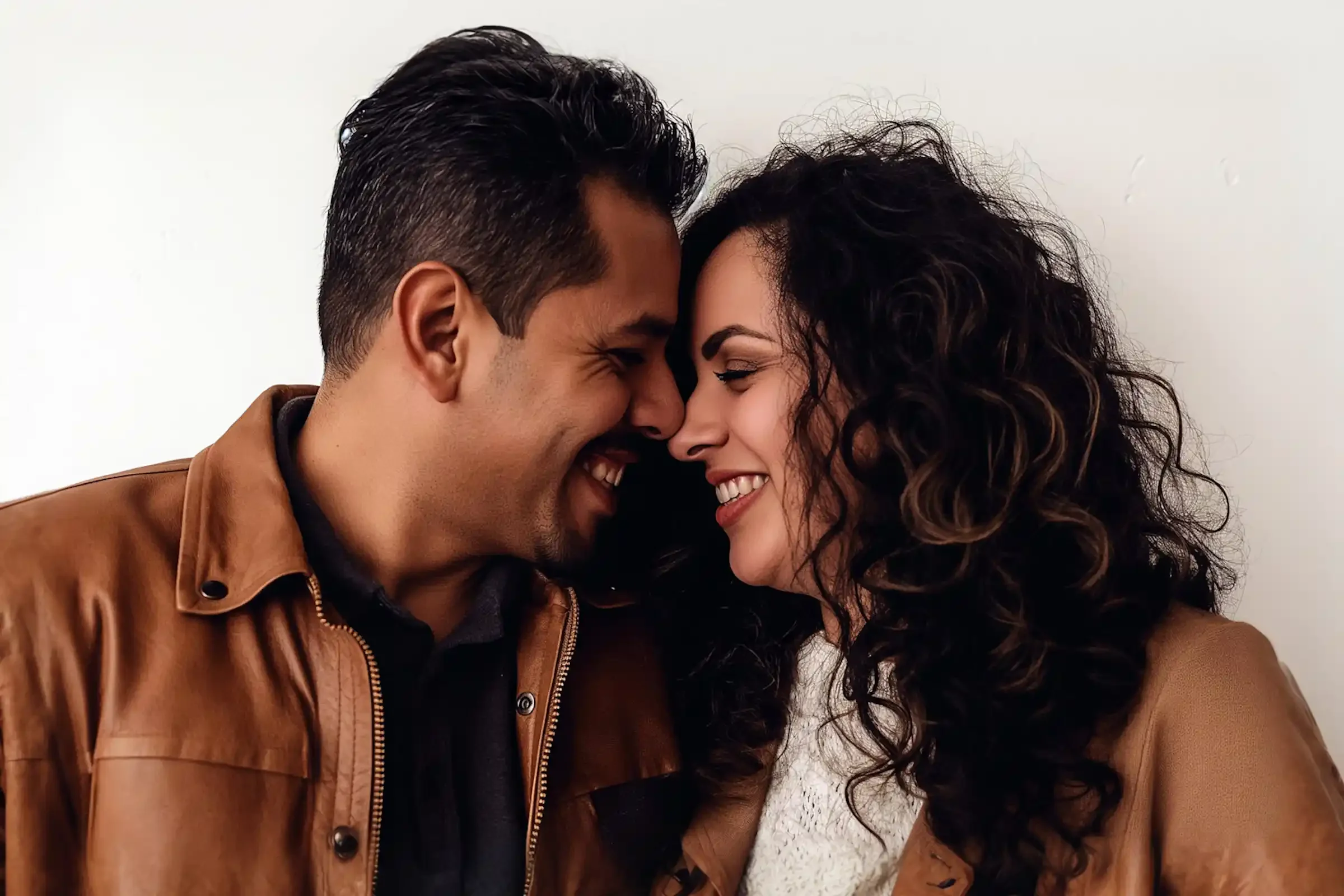 A man and woman stand close together, smiling and touching noses. Both wear brown jackets and have dark, wavy hair. The background is plain white, creating a warm and intimate atmosphere.