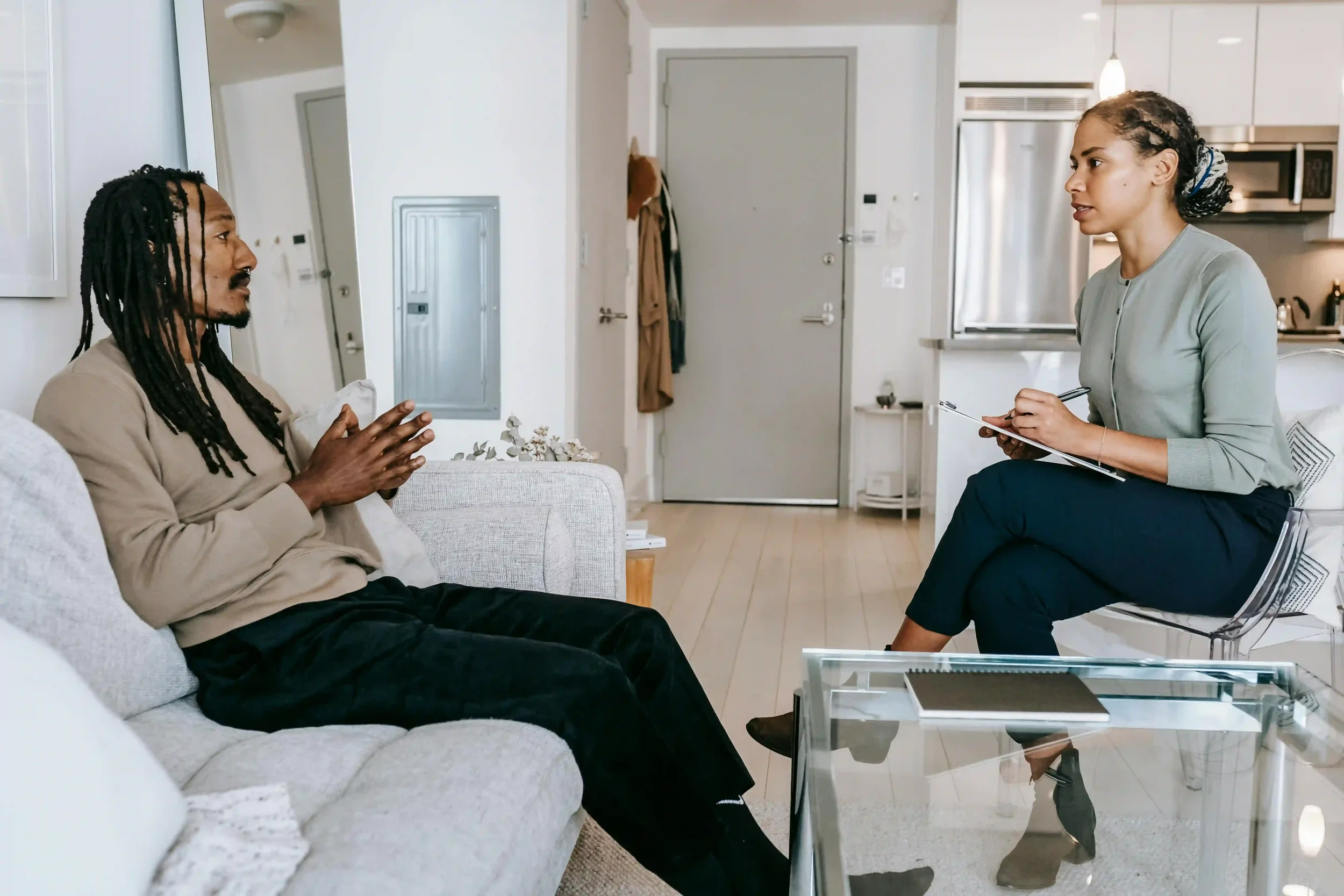 A man sits on a couch talking while a woman with a notepad listens attentively, suggesting a therapy or counseling session in a modern office..