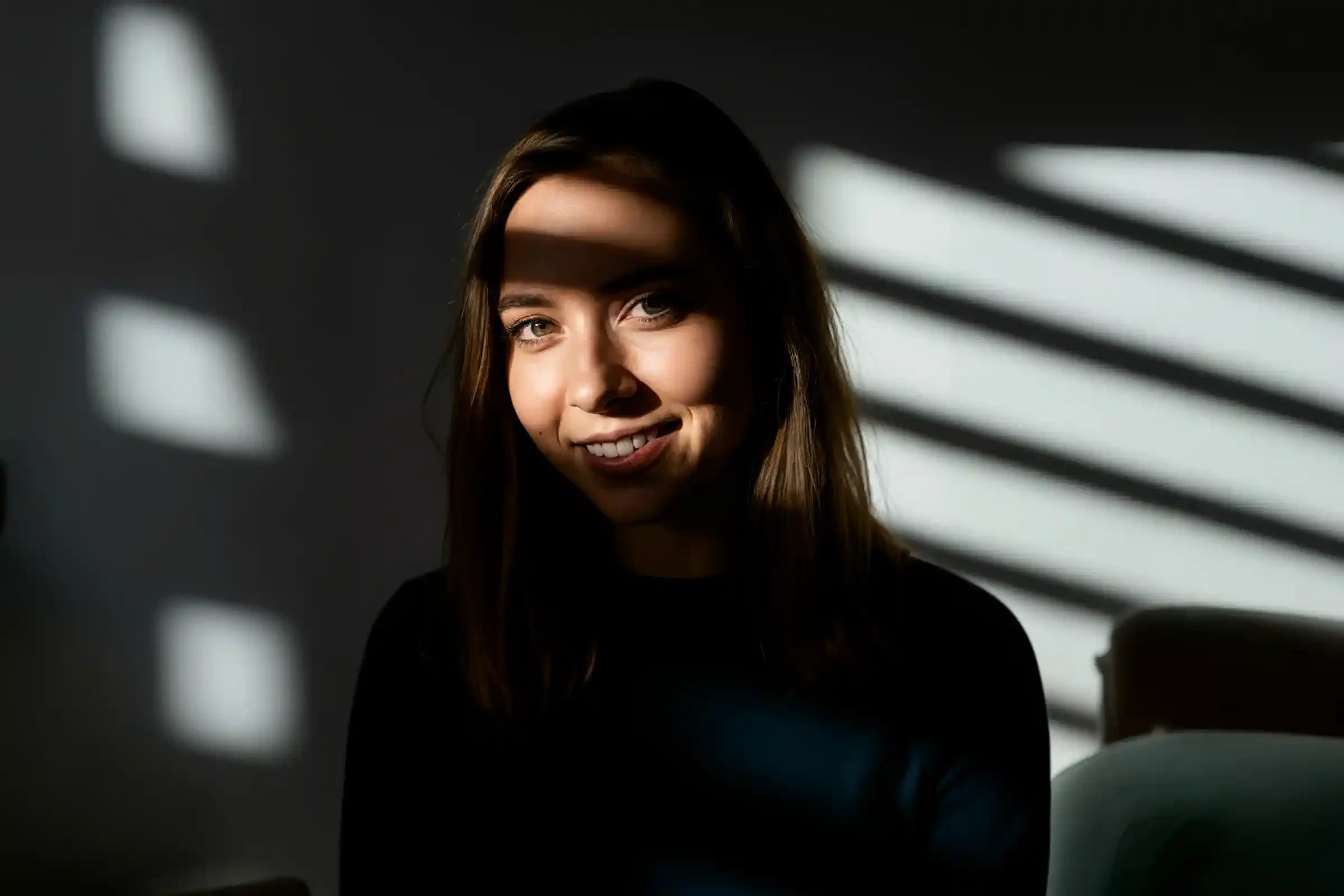 A woman with long brown hair smiles at the camera, sitting in a dimly lit room with striped sunlight and shadow patterns cast across her face and background.