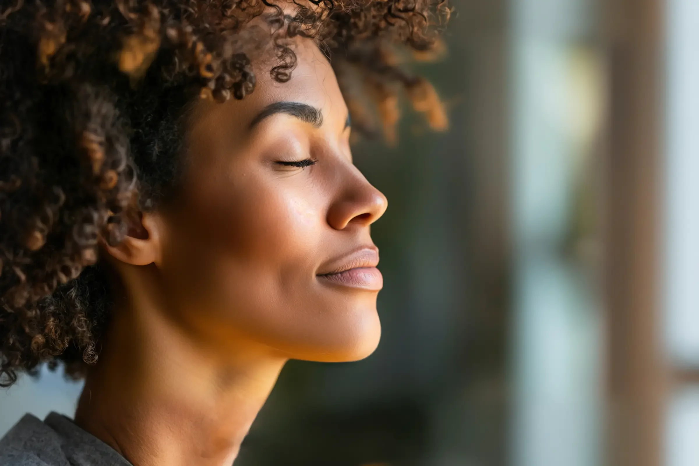 A close-up of a woman with curly hair and closed eyes, smiling softly and appearing calm and relaxed in a softly lit indoor setting.