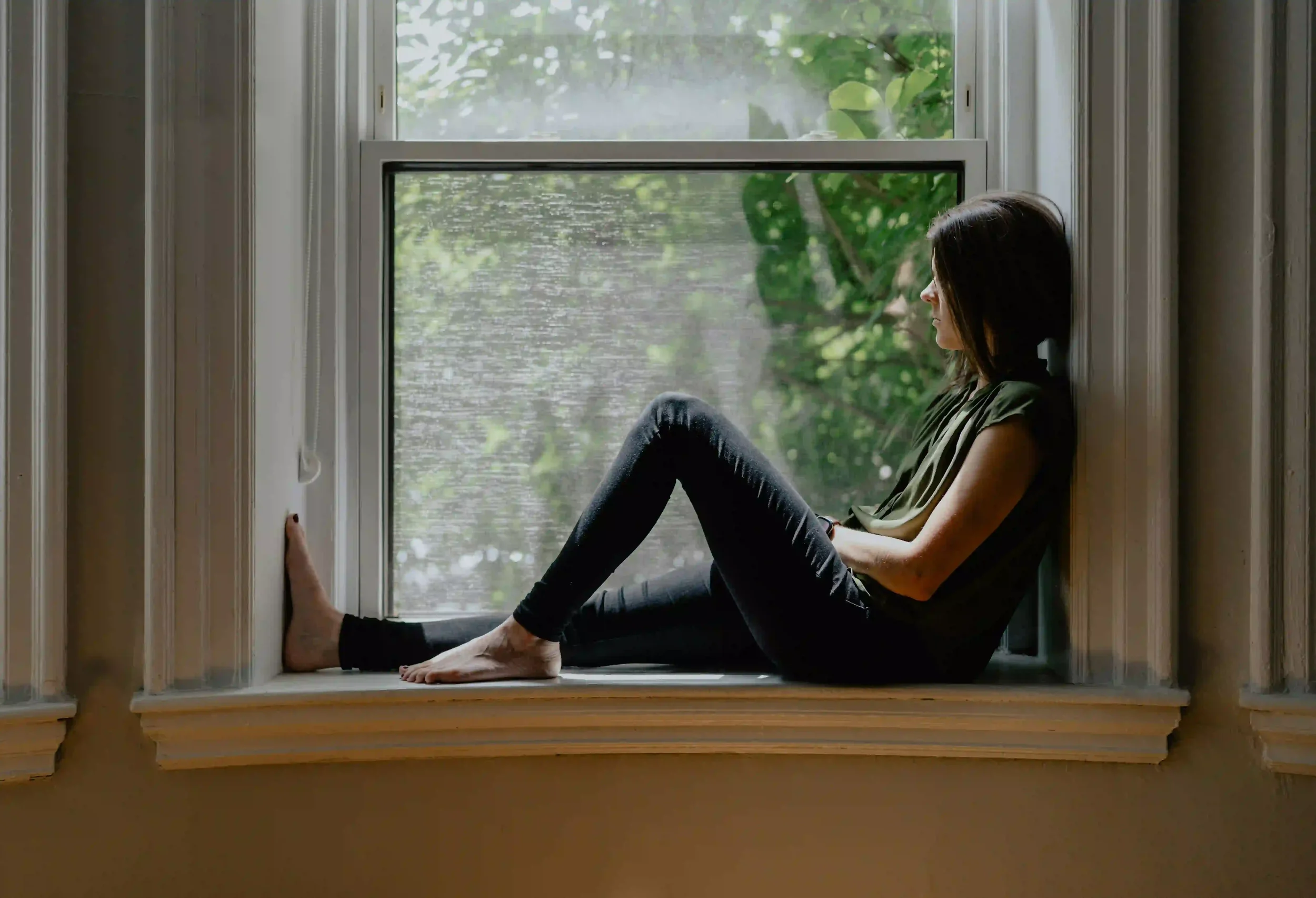 A woman sitting on a wide windowsill, looking out the window.