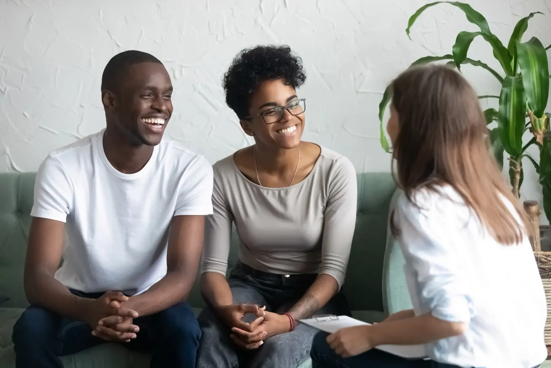 A smiling couple sits on a couch, engaged in a friendly conversation with a woman holding a clipboard, suggesting a counseling or therapy session in a comfortable, well-lit room with plants.
