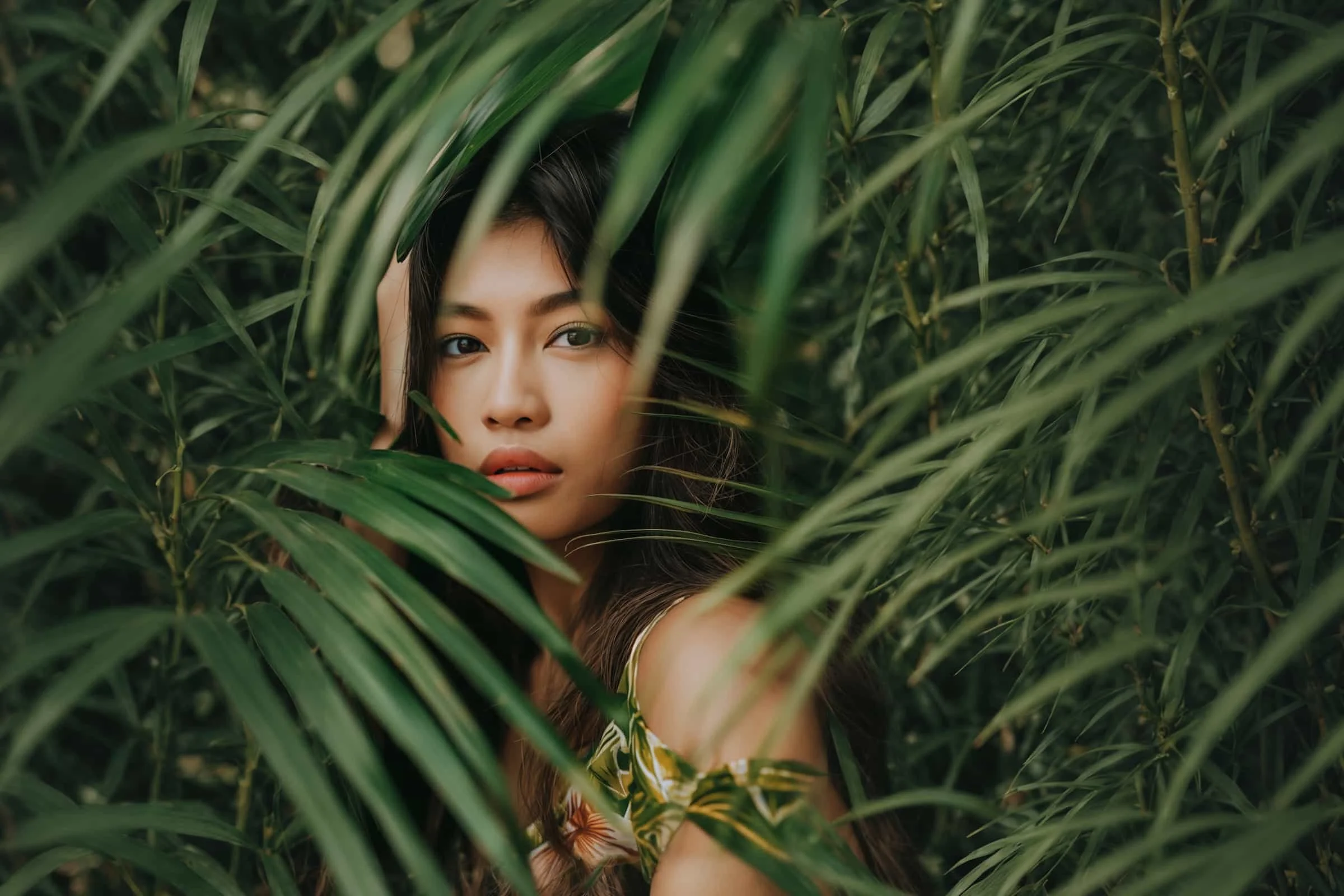 A woman with long dark hair looks through green tropical leaves, partially obscuring her face. She has a calm expression and wears a patterned outfit, blending with the lush foliage surrounding her.