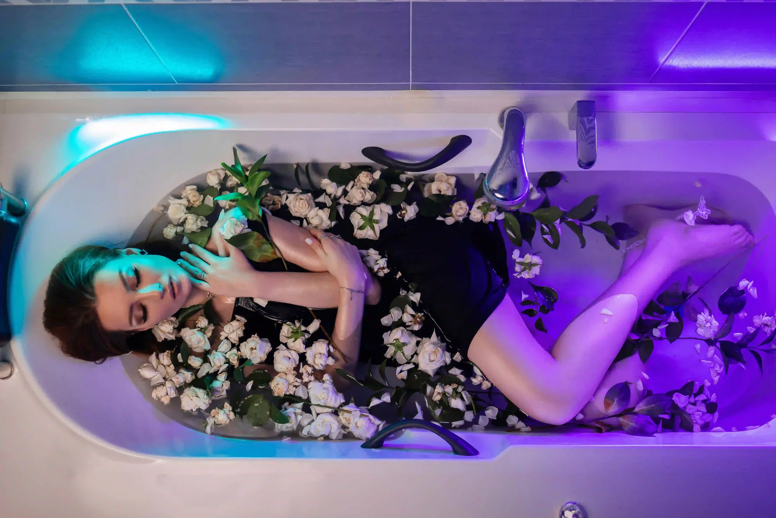 A woman in a black dress lies in a bathtub filled with white flowers and green leaves, eyes closed and hugging some flowers, under soft blue and purple lighting.