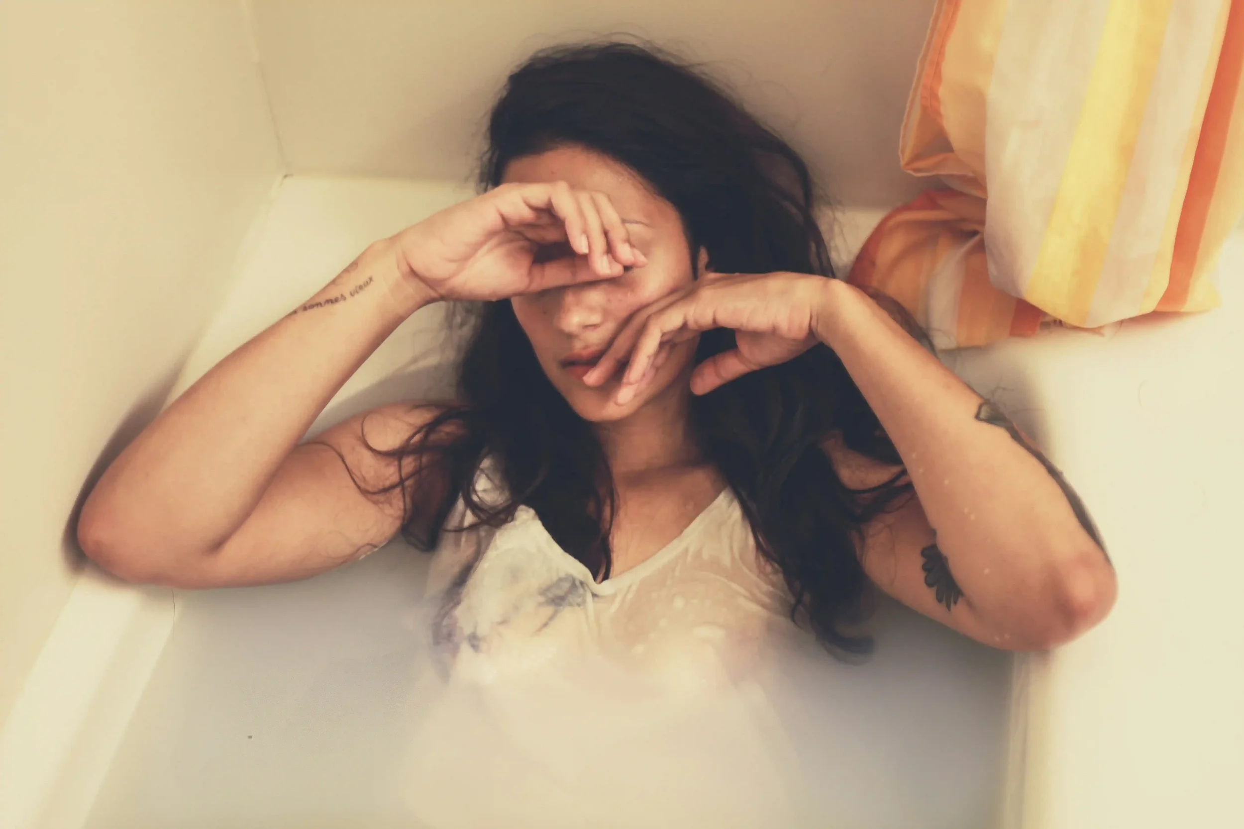 A woman with long dark hair lies in a bathtub filled with milky water, wearing a white shirt, covering her face with her hands. A yellow and orange towel hangs on the wall nearby.