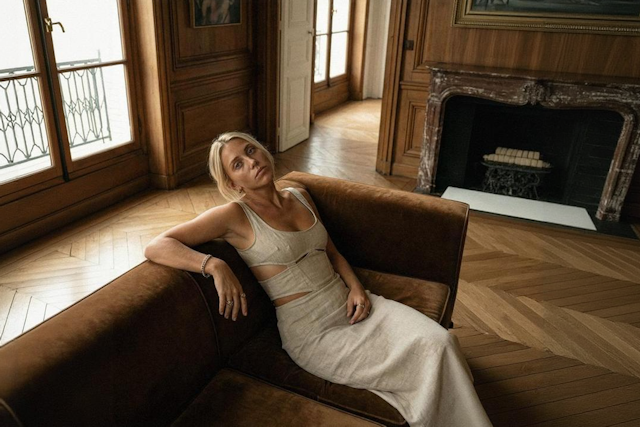 Woman reclining on a brown leather couch in a warm, sunlit room with wood floors and a fireplace, appearing distant and introspective in a moment that suggests emotional flashbacks.