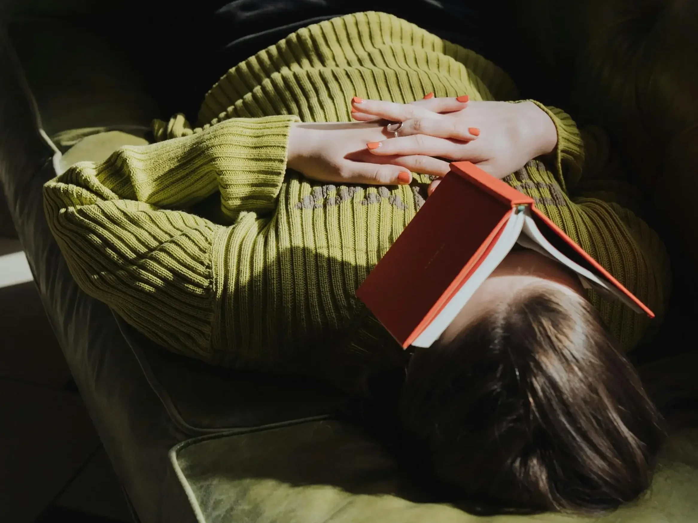 A woman lying on a couch with an open red book resting over their face.