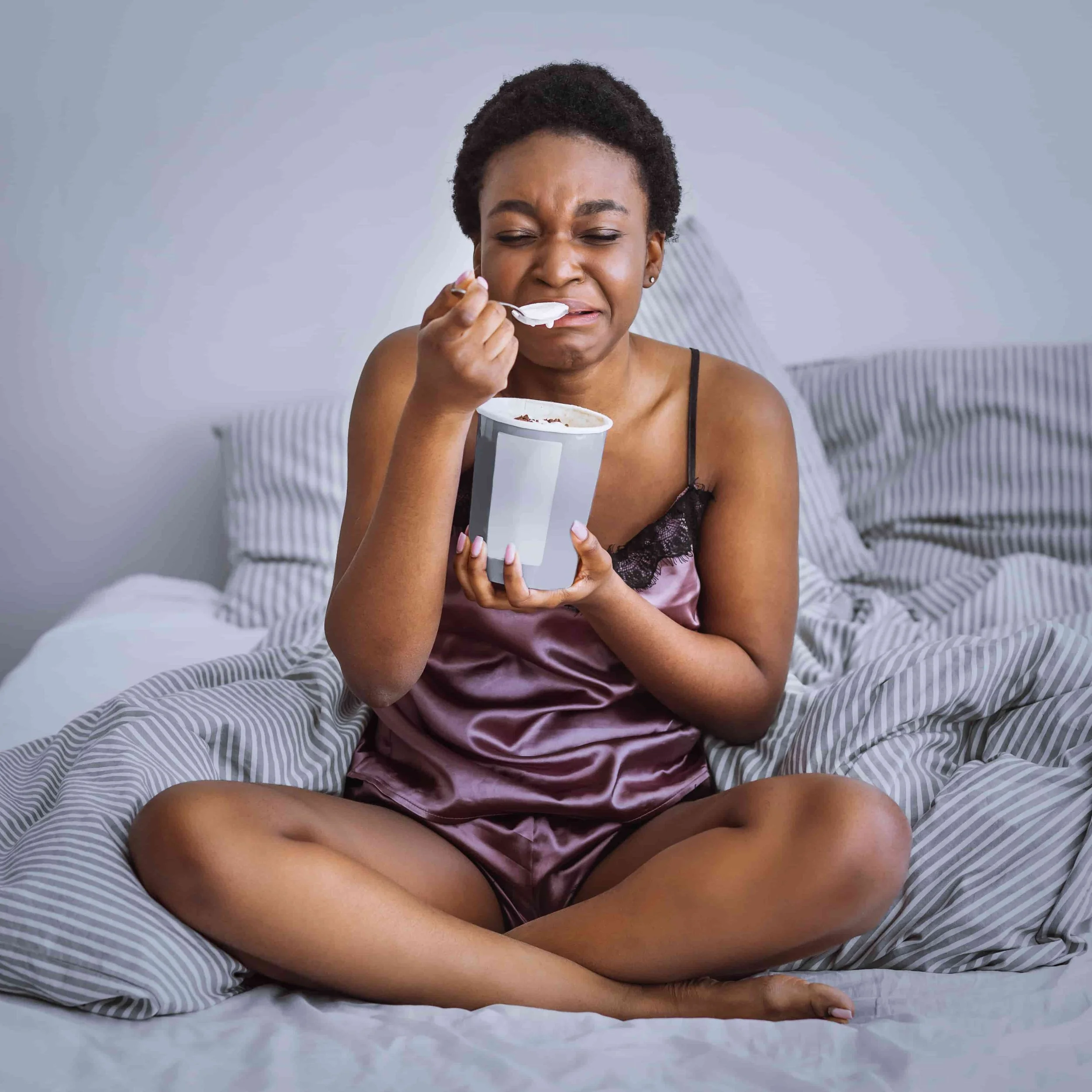 A woman in satin pajamas sits cross-legged on a bed, holding a tub of ice cream and eating with a spoon, while appearing sad and about to cry.