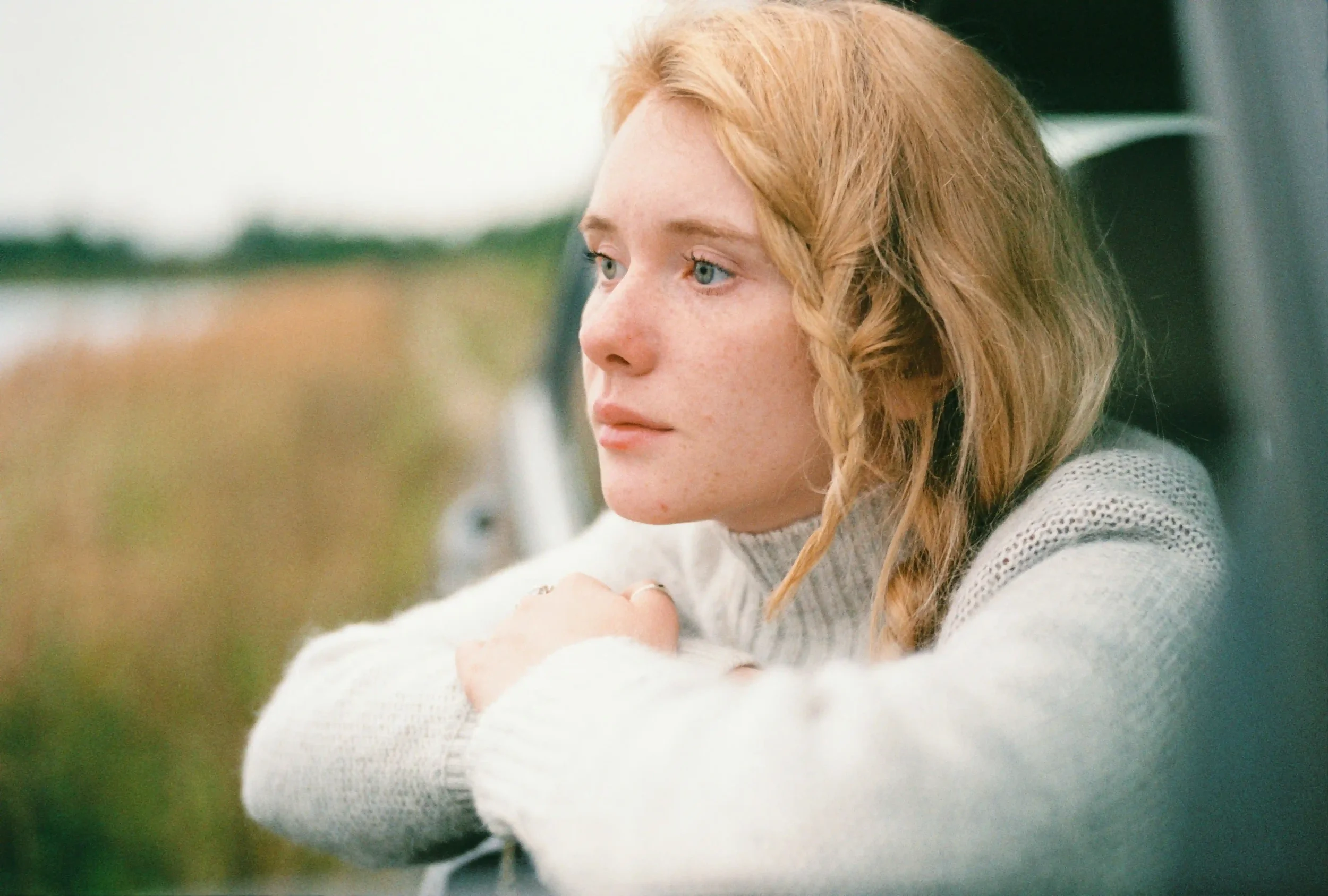 A person with long blonde hair and a braid leans out of a car window, resting their arms and looking thoughtfully into the distance. They wear a light sweater. The background is blurred with greenery.