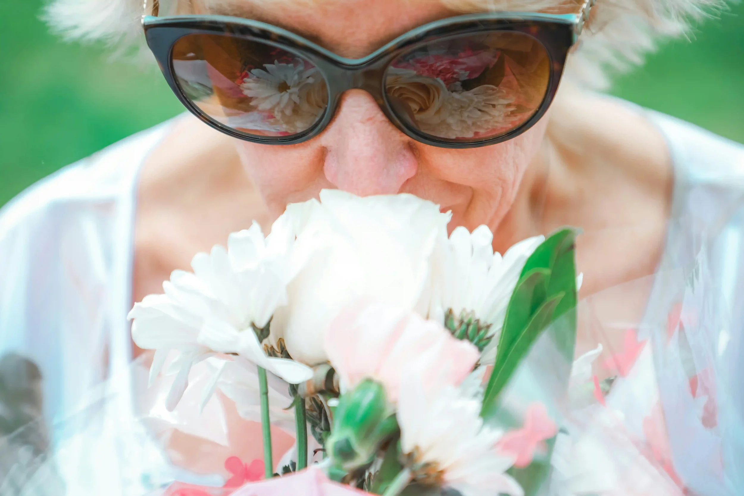An older woman wearing sunglasses is smelling a bouquet of white and pale pink flowers. The image is close-up, focusing on her face and the flowers, with a green background out of focus.
