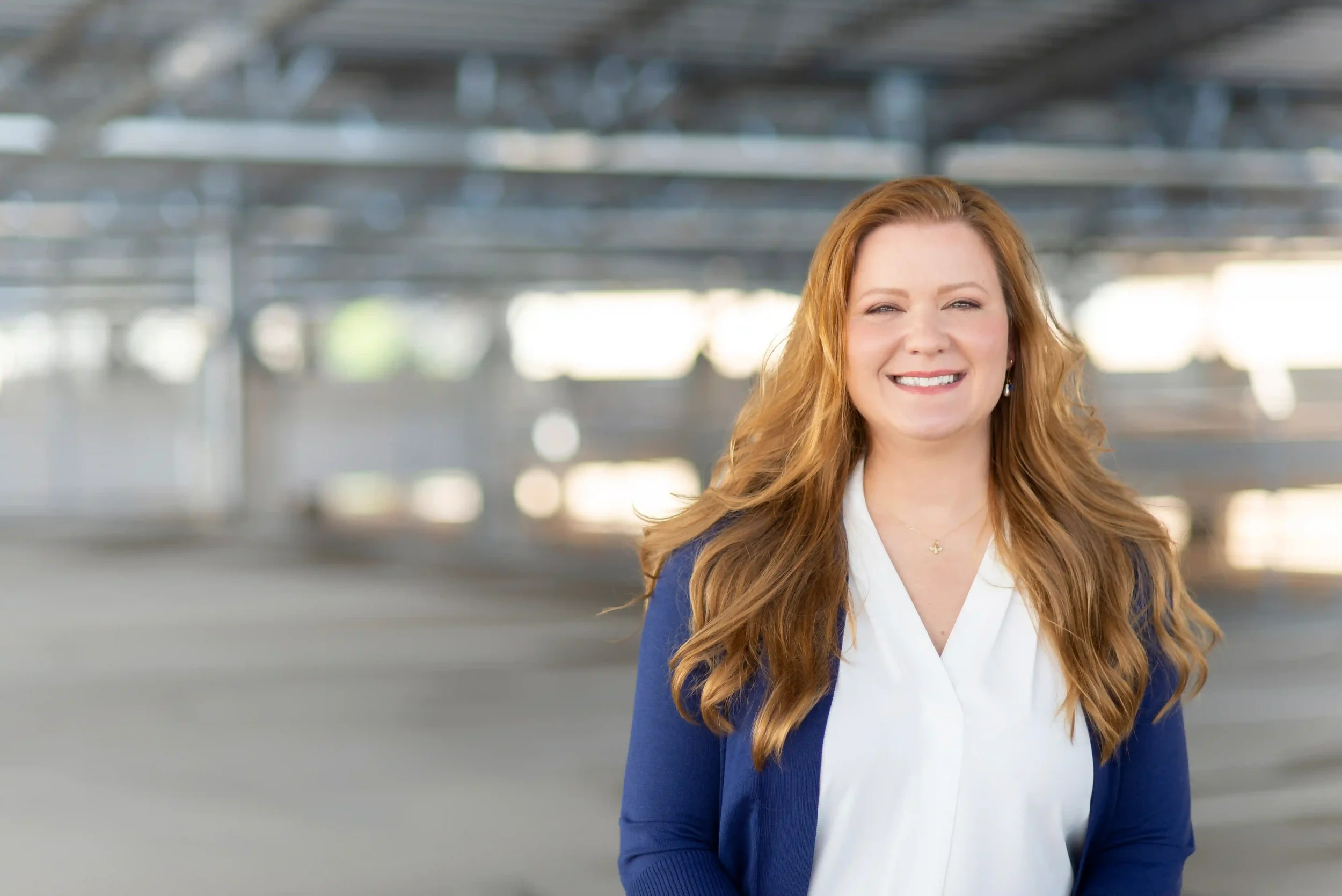 A woman with long red hair, wearing a white blouse and blue cardigan, stands smiling in a bright, empty indoor space with a blurred background.