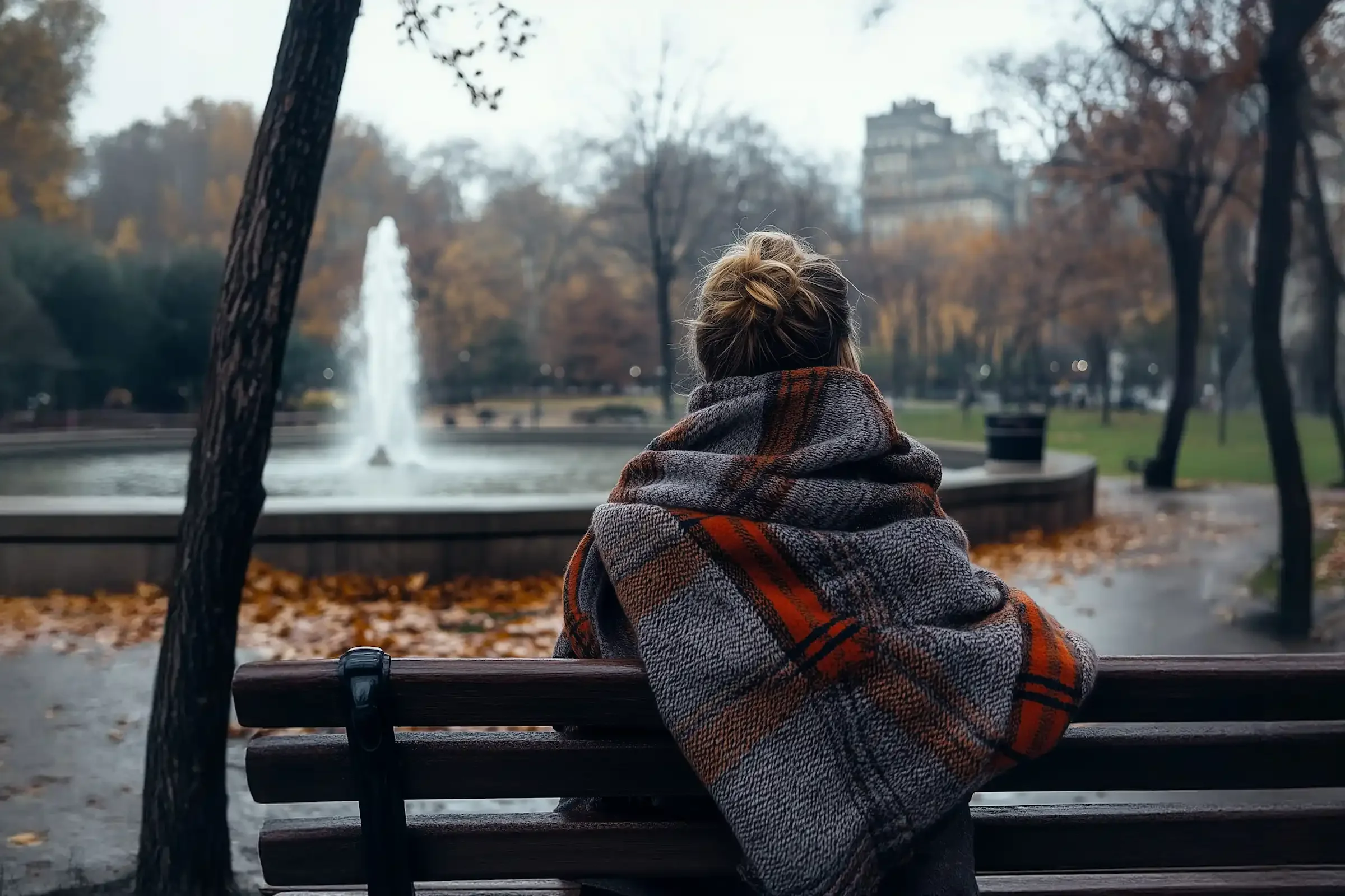 A person wrapped in a plaid blanket sits on a park bench, facing a fountain and autumn trees on a cloudy day.