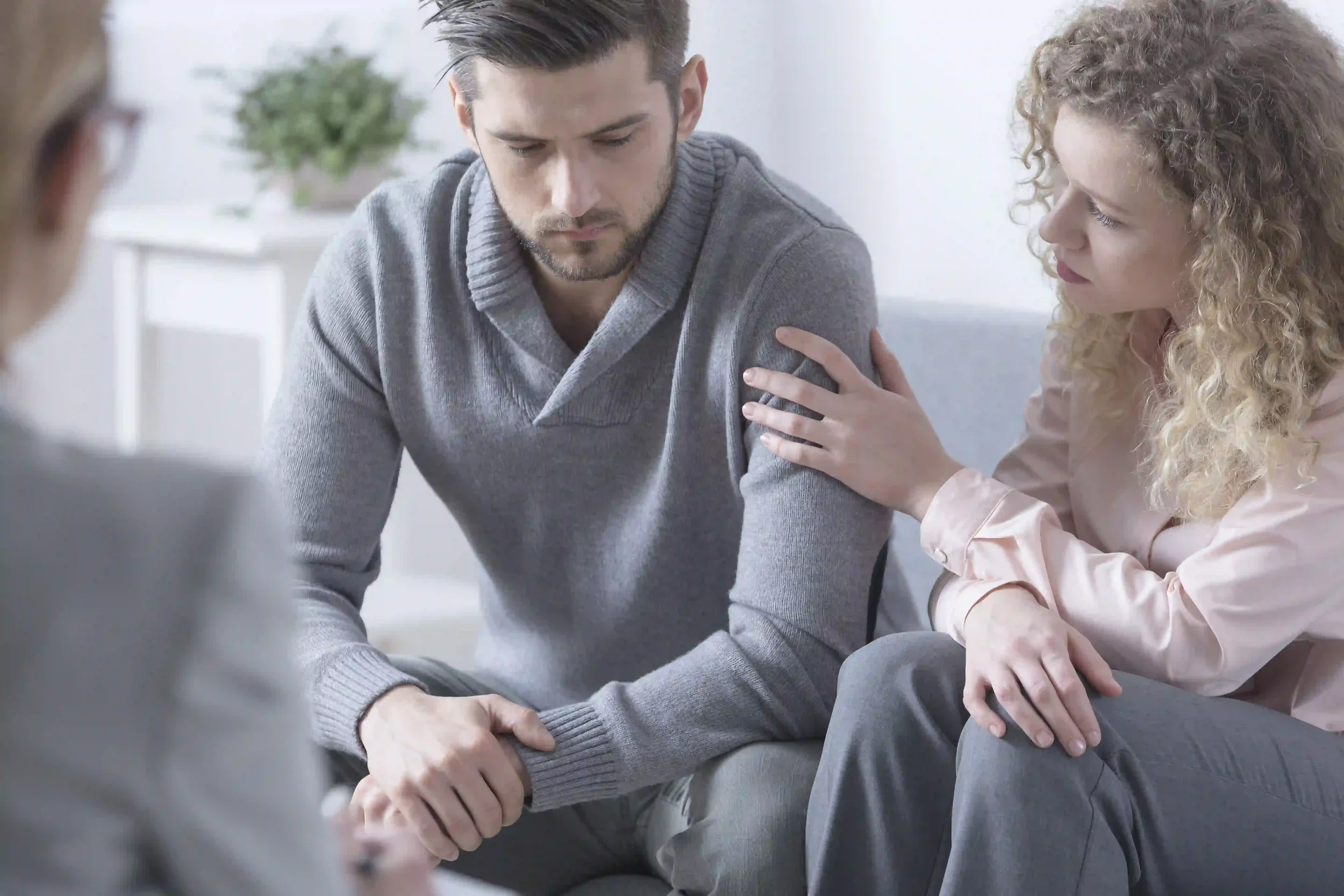 A woman comforts a sad man by gently touching his arm during a counseling session, while another person sits nearby, partially visible in the foreground.
