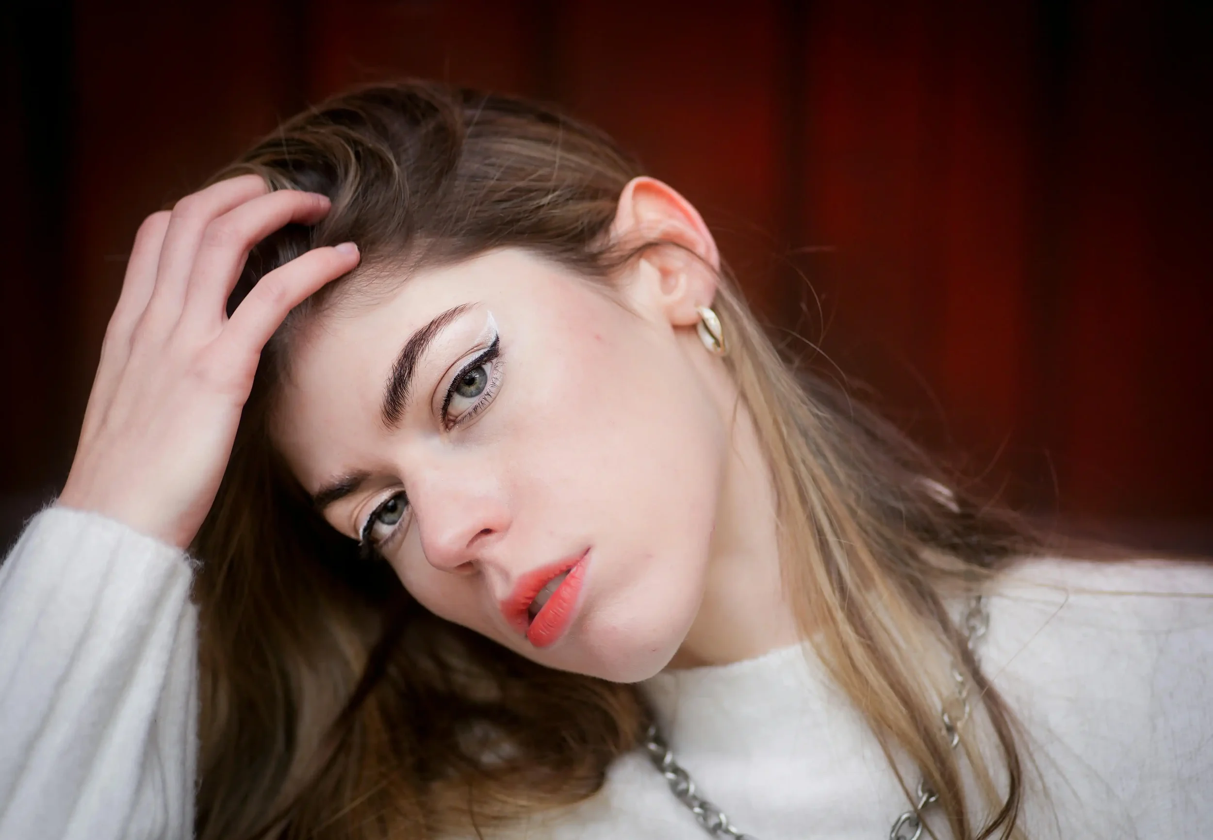 A person with long light brown hair, bold eyeliner, red lipstick, and a white top looks at the camera while touching their head. The background is blurred and dark red.