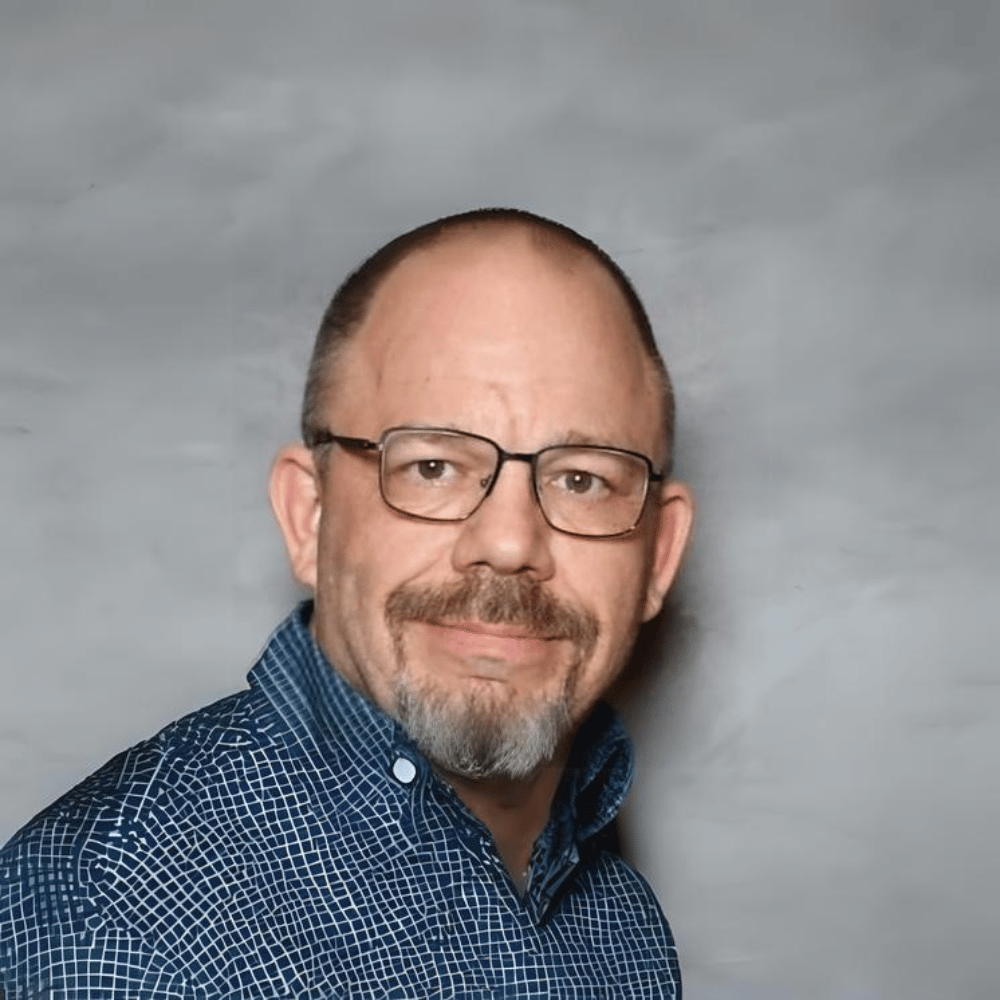A middle-aged man with glasses, short hair, and a goatee, wearing a blue patterned shirt smiling at the camera against a gray background.