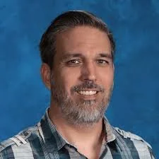 A man with dark hair and a beard, wearing a plaid shirt, smiling against a blue background.