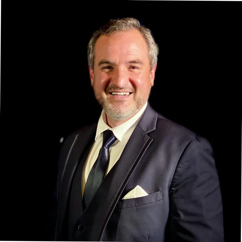 A smiling man in a formal suit, white shirt, and dark tie posing against a black background.