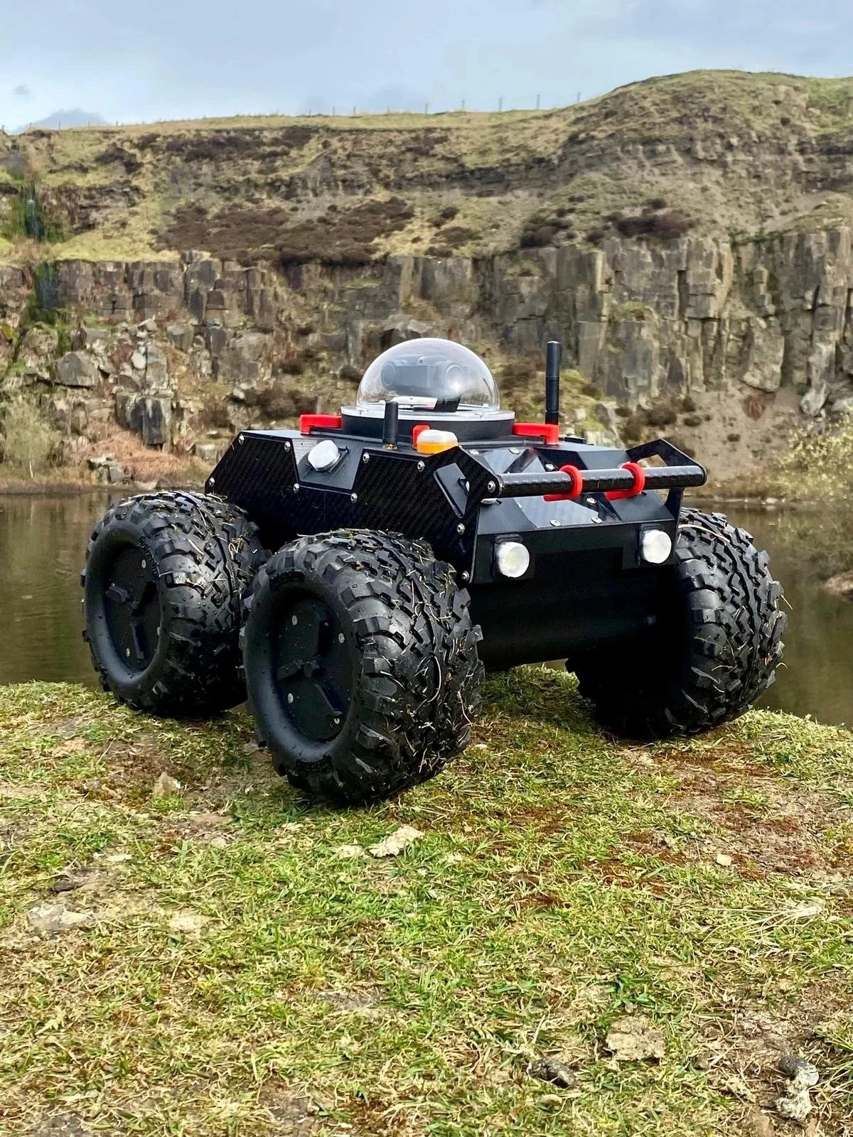A small robotic vehicle with large off-road tires, black body, and a clear dome on top, outside in front of a rocky hillside and a body of water.