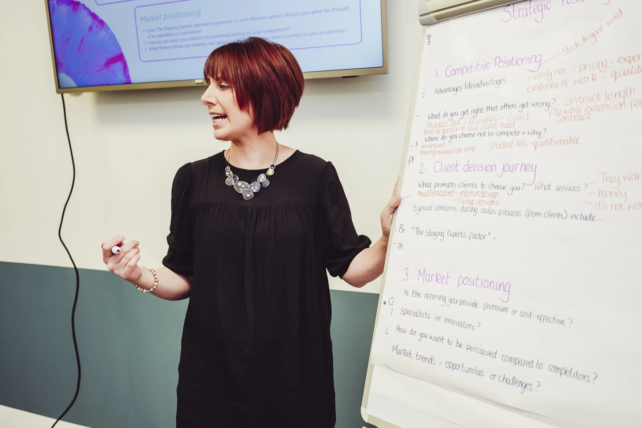 A woman with short reddish-brown hair wearing a black dress and a chunky necklace is giving a presentation. She is holding a marker in her right hand and pointing to a whiteboard filled with handwritten notes on marketing topics. There is a large monitor behind her displaying a slide related to market positioning.