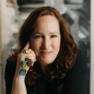 Close-up portrait of a woman with red hair, smiling gently at the camera, with her hand resting on her face in a thoughtful pose, in an indoor setting with blurred background.