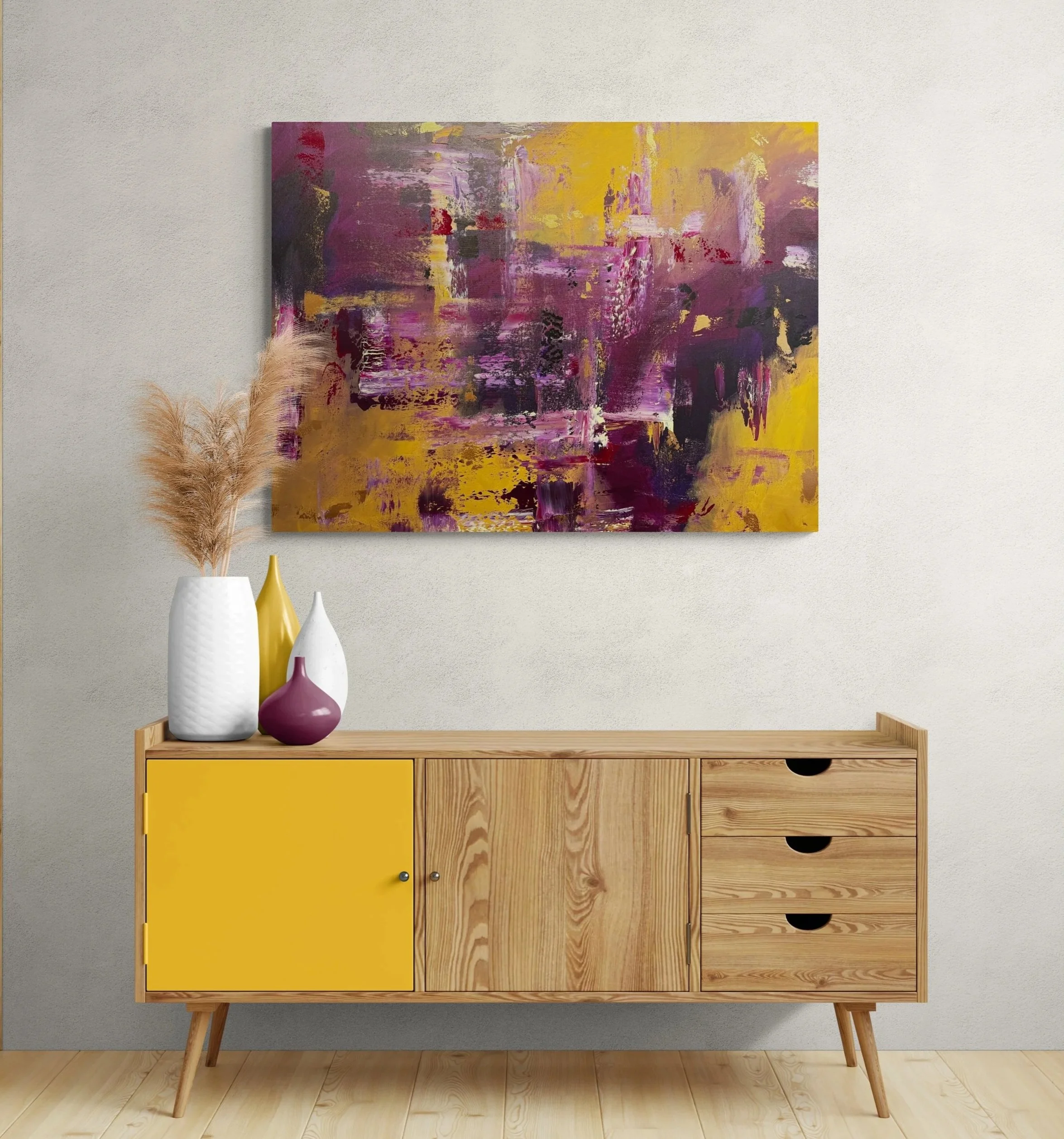 This image shows a modern wooden sideboard with yellow and natural wood drawers, decorated with vases, against a light-colored wall with an abstract painting in shades of purple, yellow, and black. There are three vases, two white and one yellow, on top of the sideboard, and a large abstract painting hangs above it.