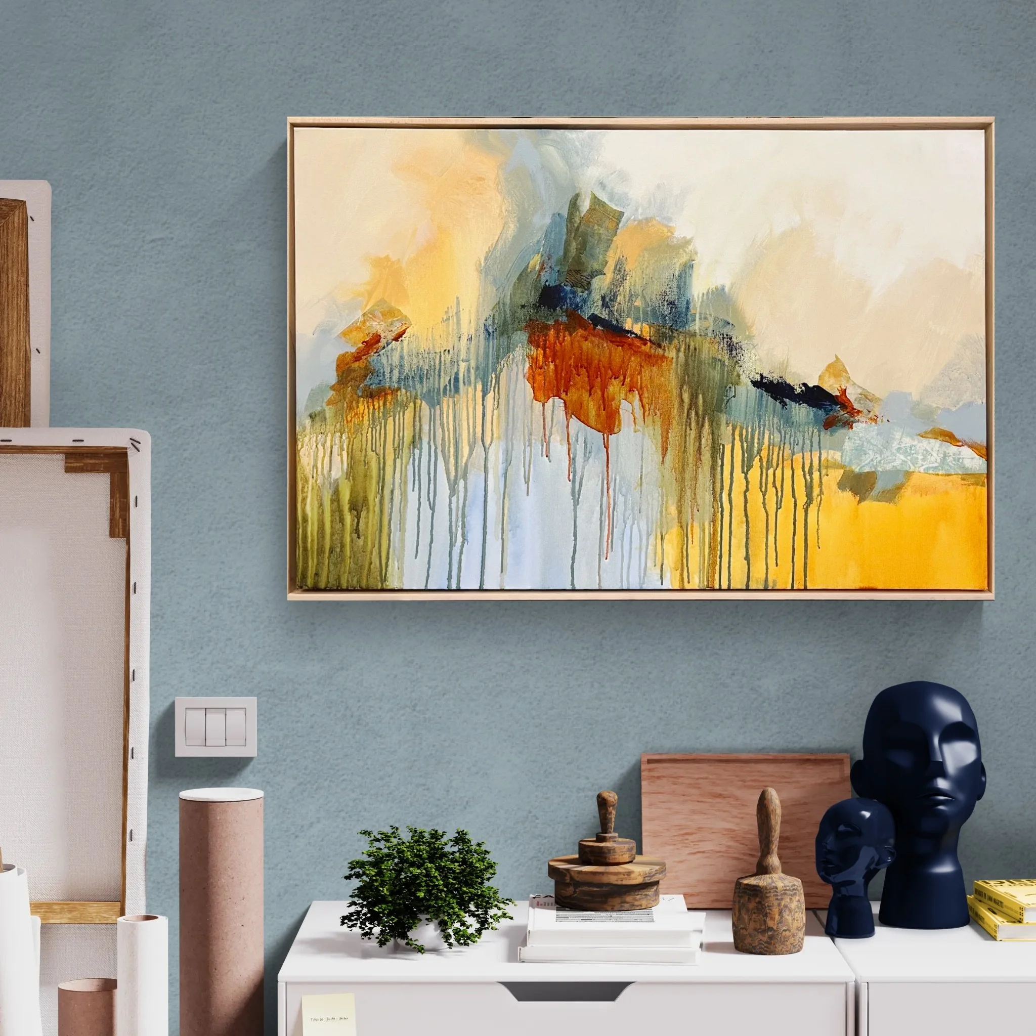 Abstract mountain landscape painting with yellow, orange, blue, and green hues hanging on a blue wall above a white sideboard. The sideboard has decorative items including a small green plant, wooden sculptures, black face bust sculptures, and stacks of books.