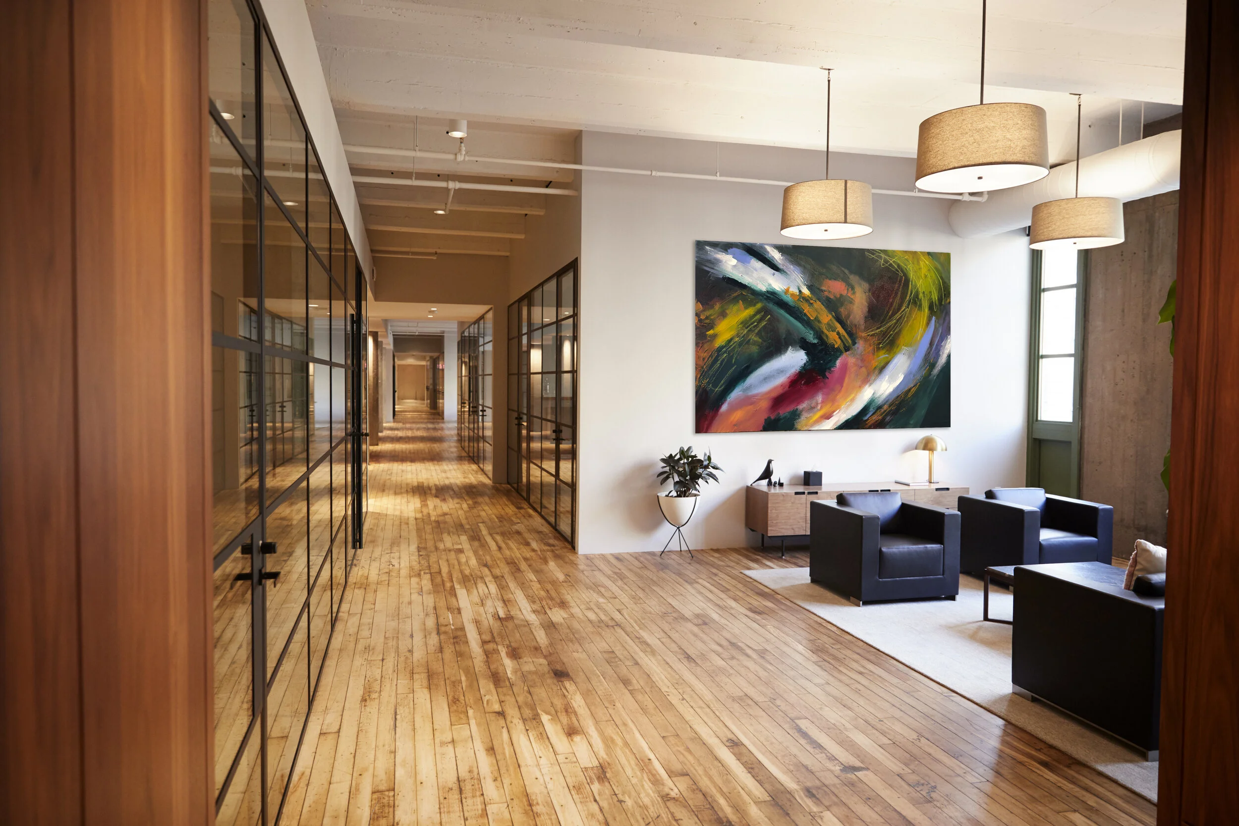 Modern office lounge area with black armchairs, a wooden coffee table, a large abstract painting, a potted plant, and hanging ceiling lights.