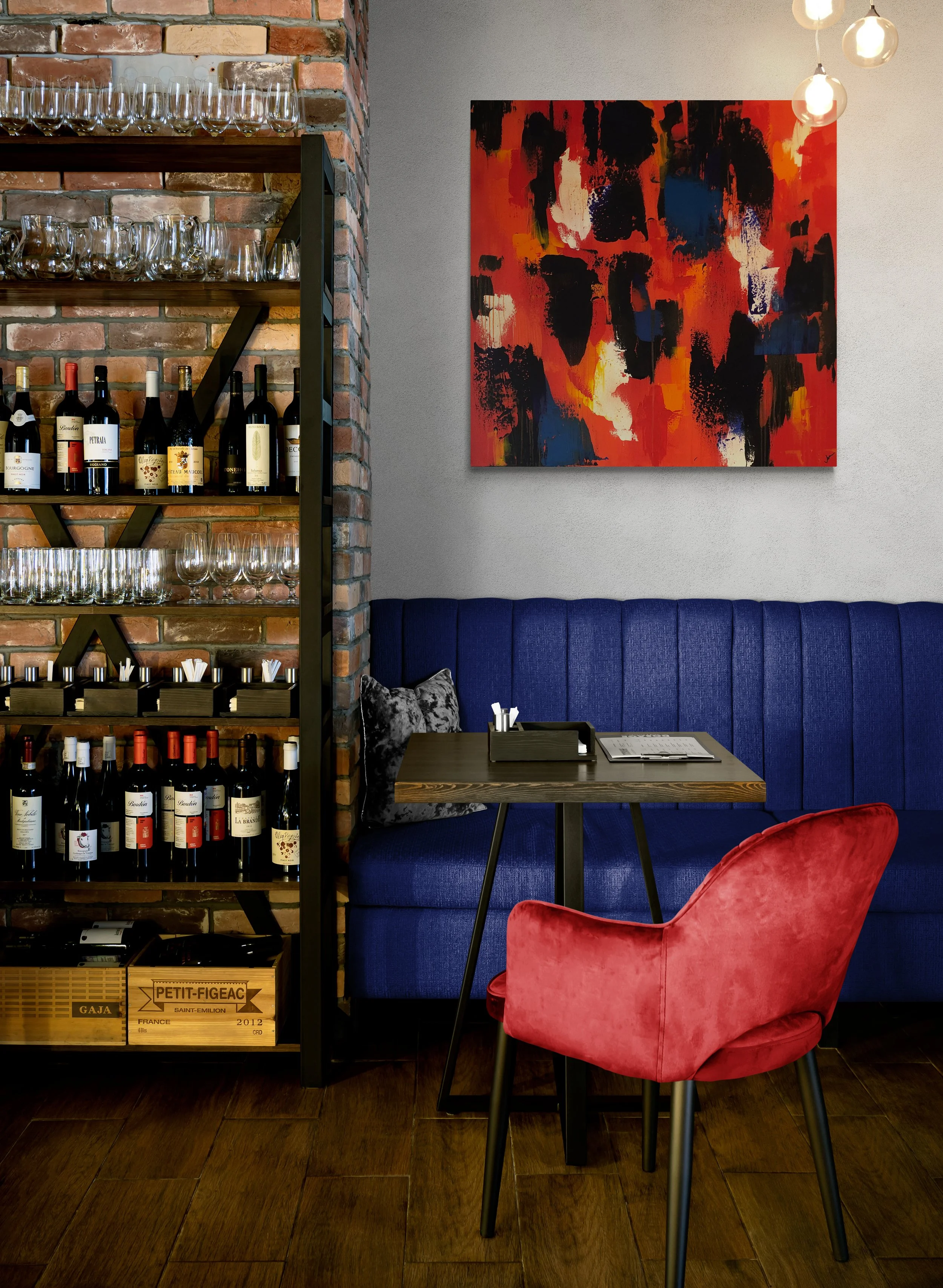 A cozy restaurant corner featuring a brick wall with shelves holding wine bottles and glasses, a colorful abstract painting on a gray wall, a dark wood table with a black tray and menu, a plush navy blue banquette. and a pink velvet chair with wood l