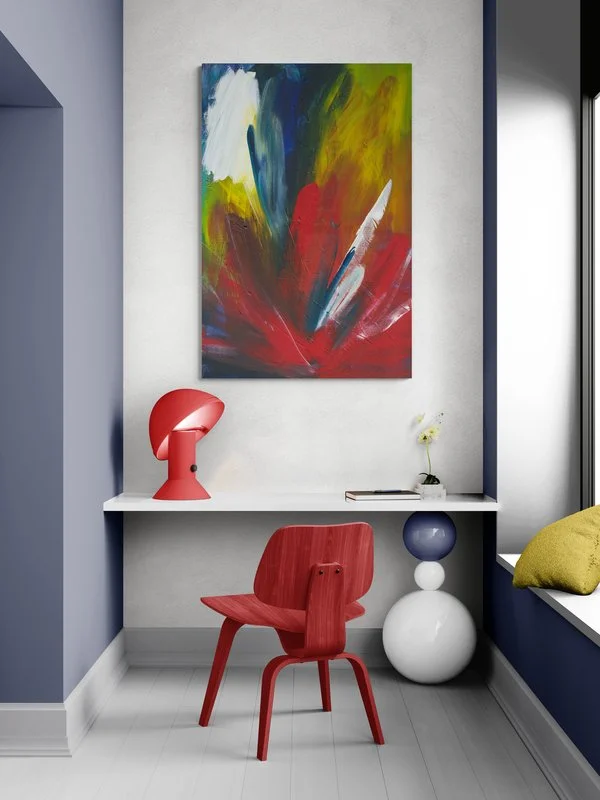 Colorful abstract painting hanging on a white wall above a workspace with a red chair, red desk lamp, a small vase with a flower, and stacked objects on a small shelf. There is a window with a yellow pillow on the window seat.