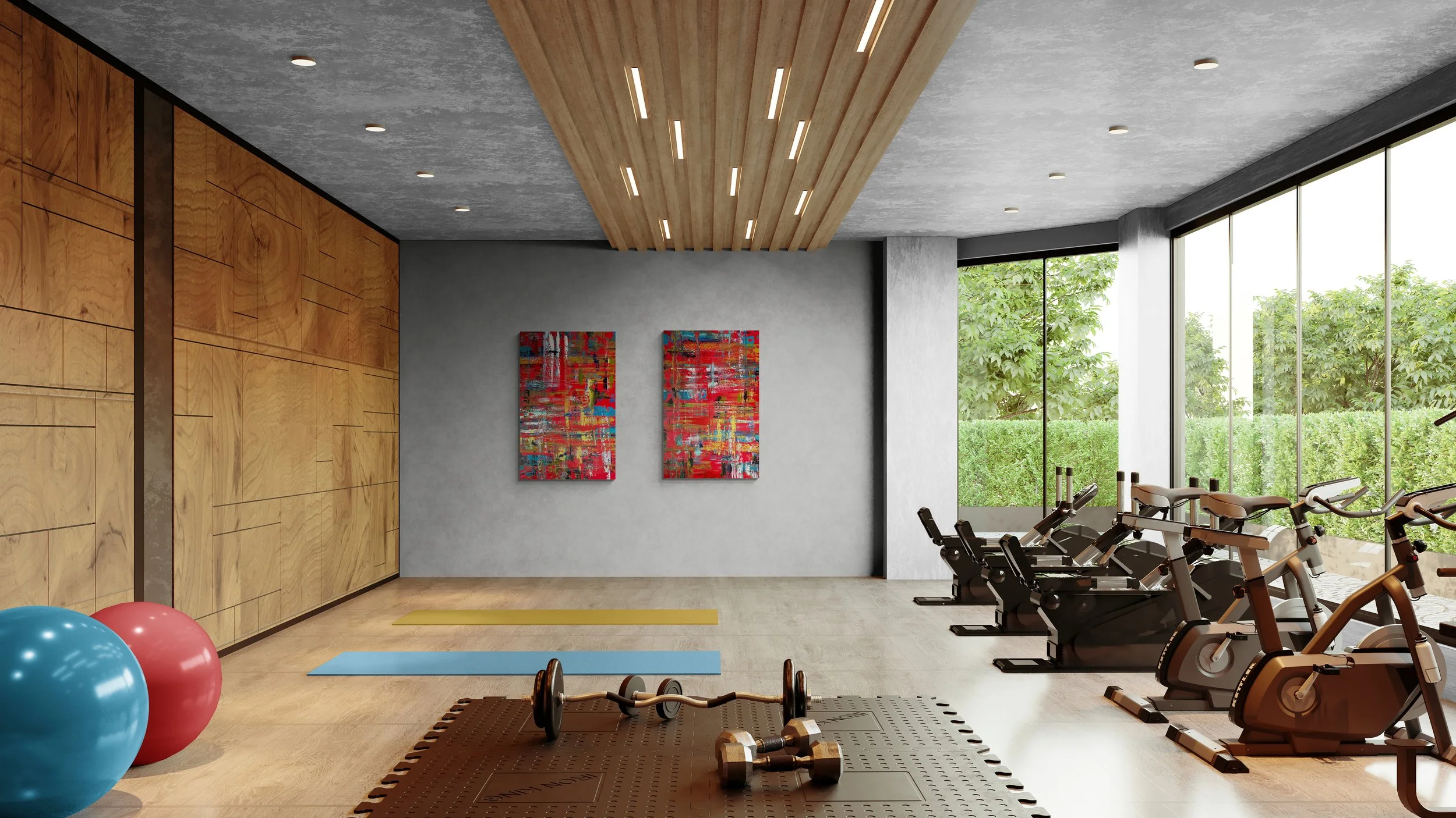 A modern gym with large windows overlooking greenery, featuring exercise bikes, yoga mats, stability balls, dumbbells, and a wooden panel wall with two colorful abstract paintings.