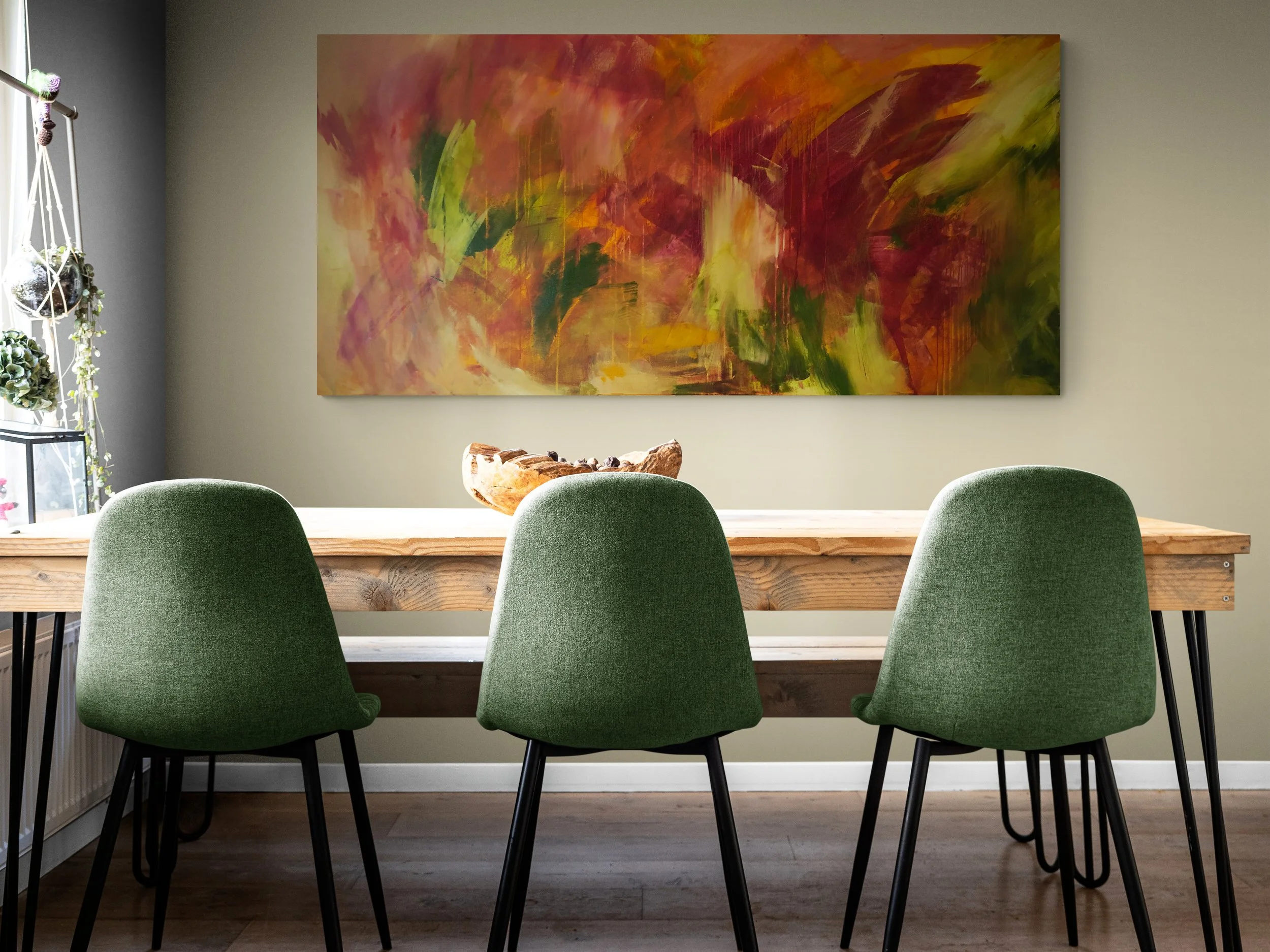 Dining room with a wooden table, three green chairs, a colorful abstract painting on the wall, and a window with hanging planters.