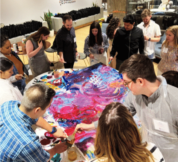 People gathered around a large canvas painting, painting and collaborating together in an art studio or workshop.