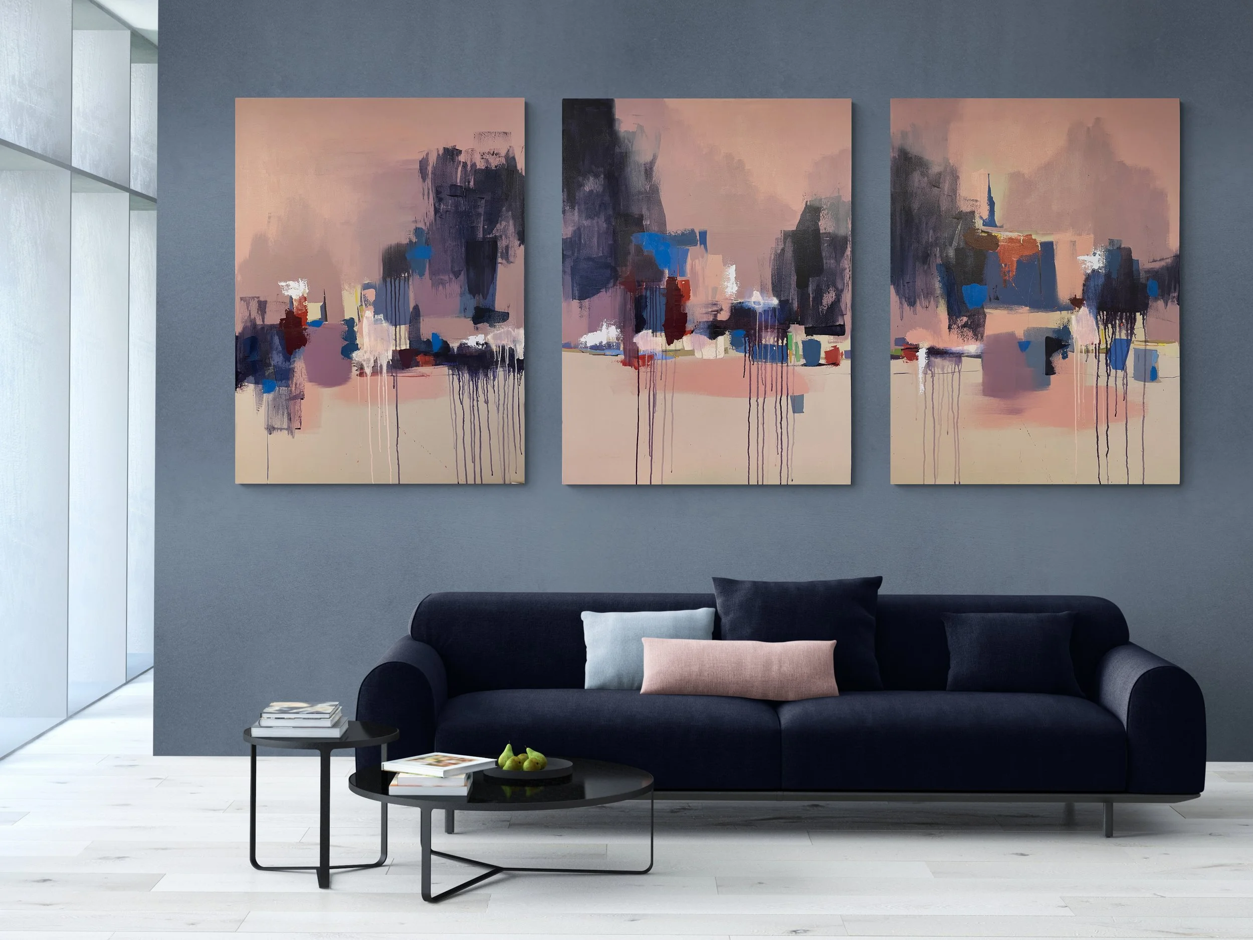 Modern living room with a dark blue sofa, three abstract cityscape paintings on a mauve wall, light-colored floor, and a small black coffee table with books and pears.