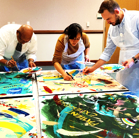 Three people painting vibrant, colorful artwork on large canvases spread on a table.